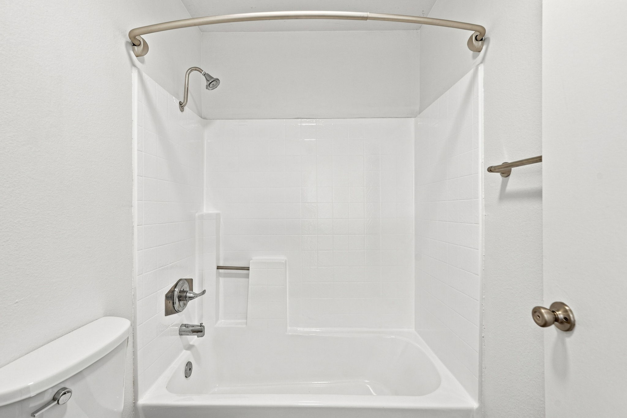 A clean, bright bathroom featuring a white bathtub with a showerhead and a curved shower rod. The tile walls are also white, creating a fresh and spacious feel. A toilet is situated to the left, with a minimalist design and a door on the right leading to another area.