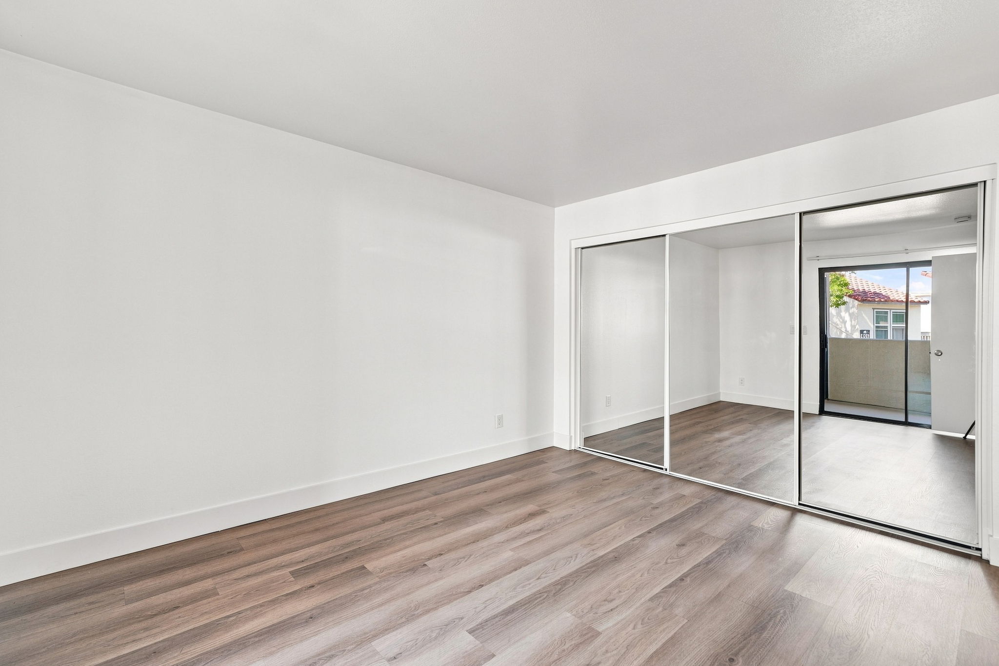 A bright, empty room with white walls and a large mirrored closet. The floor features light wood laminate, and there is a balcony door visible in the background, leading to an outdoor area. Natural light fills the space, creating an airy atmosphere.