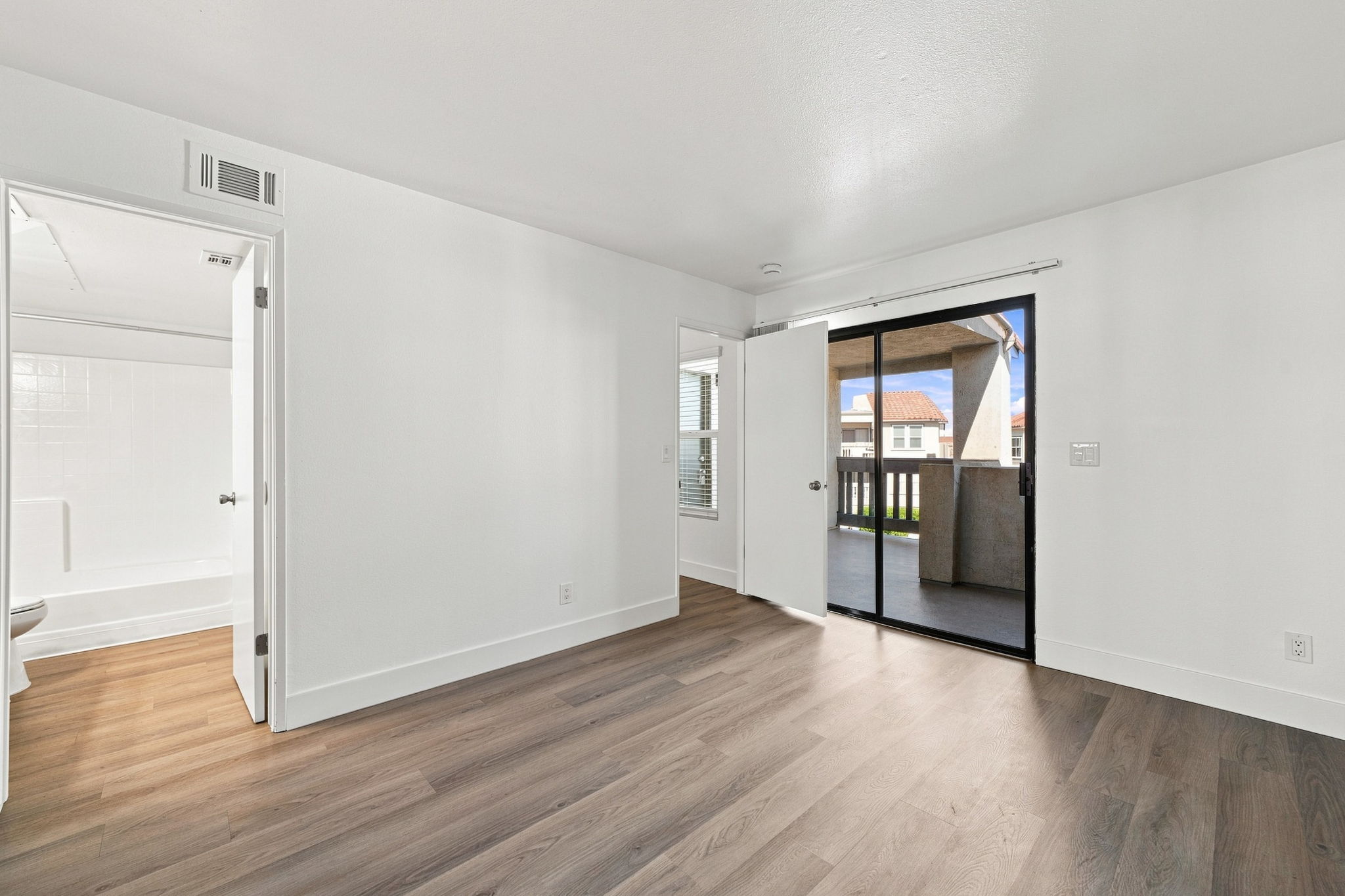 A brightly lit, empty room with light-colored walls and wood laminate flooring. There is a door leading to a balcony and an open doorway leading to a bathroom. The room has large windows with natural light coming in, creating a spacious feel.