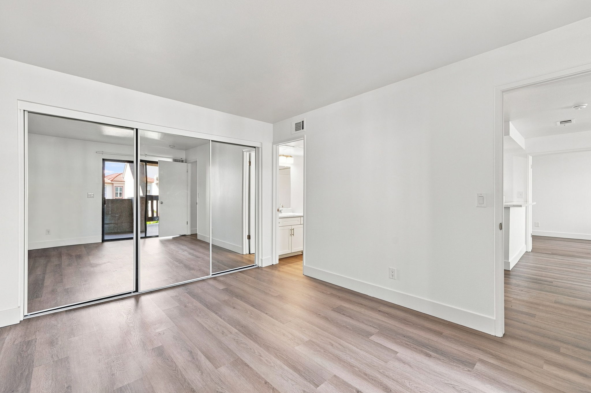 Empty room with hardwood flooring and white walls. A large mirror closet is visible on the left side, reflecting soft natural light. To the right, there's an open doorway leading to another room, and a bathroom can be seen in the background. The space appears bright and modern, suitable for furnishing.
