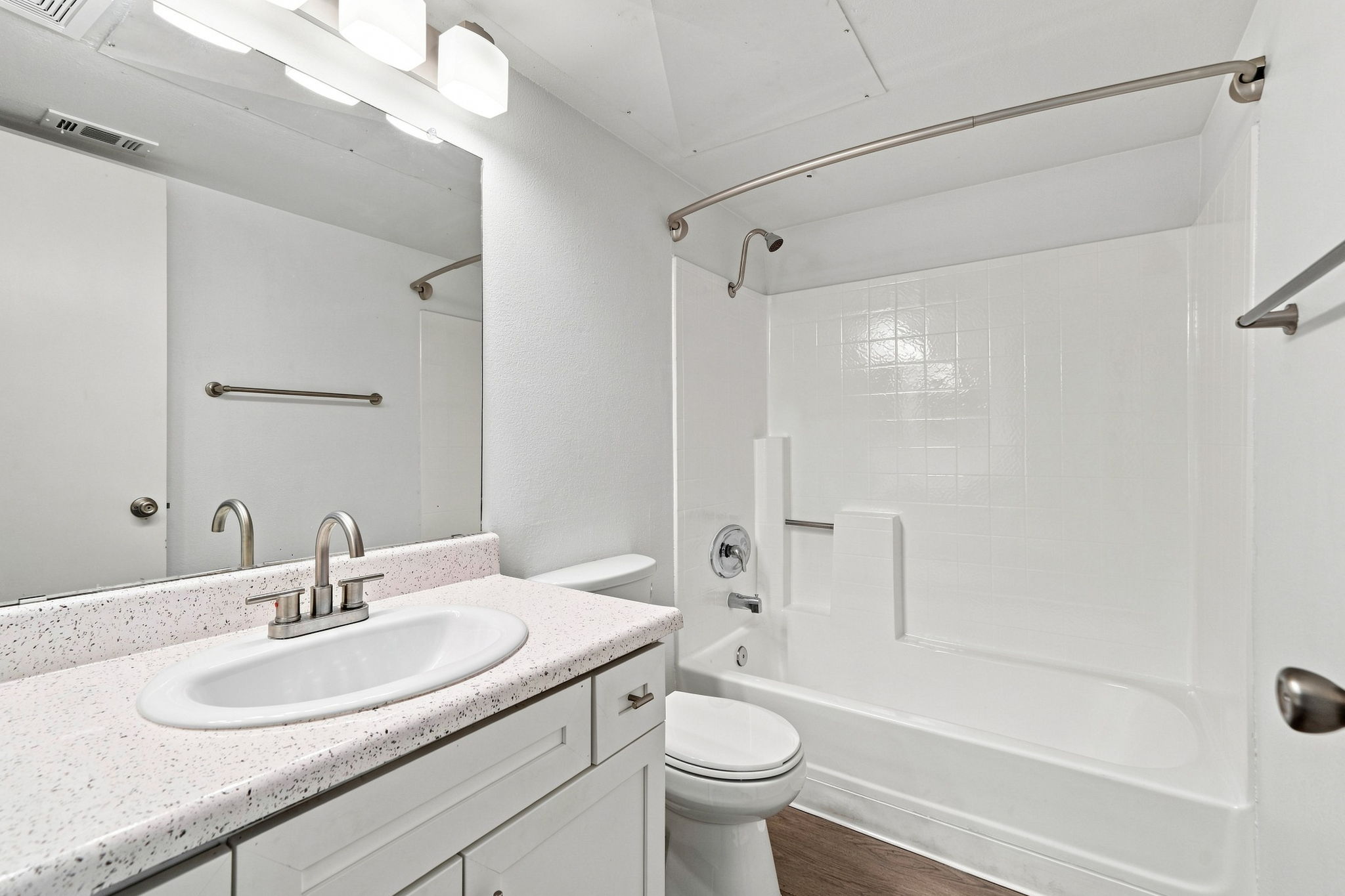 A clean and modern bathroom featuring a white sink with a single faucet, a toilet, and a bathtub with a shower. The walls are painted white, and there's a large mirror above the sink, illuminated by three light fixtures. The floor appears to be a dark wood or laminate material.