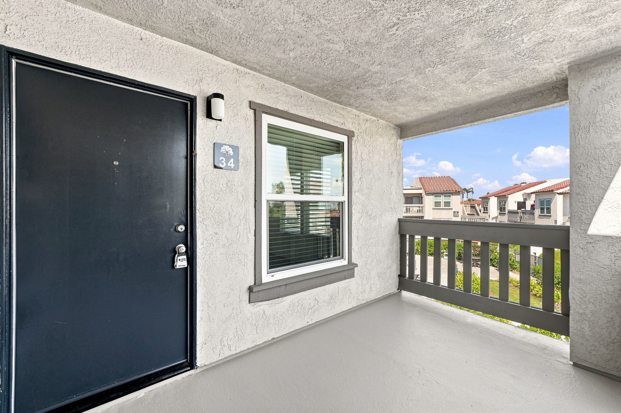 A view of a porch area with a dark blue front door marked with the number 34. There's a window with blinds to the right, and the background features a glimpse of other buildings and blue skies. The floor is light gray, and the walls are a neutral color, creating a welcoming outdoor space.