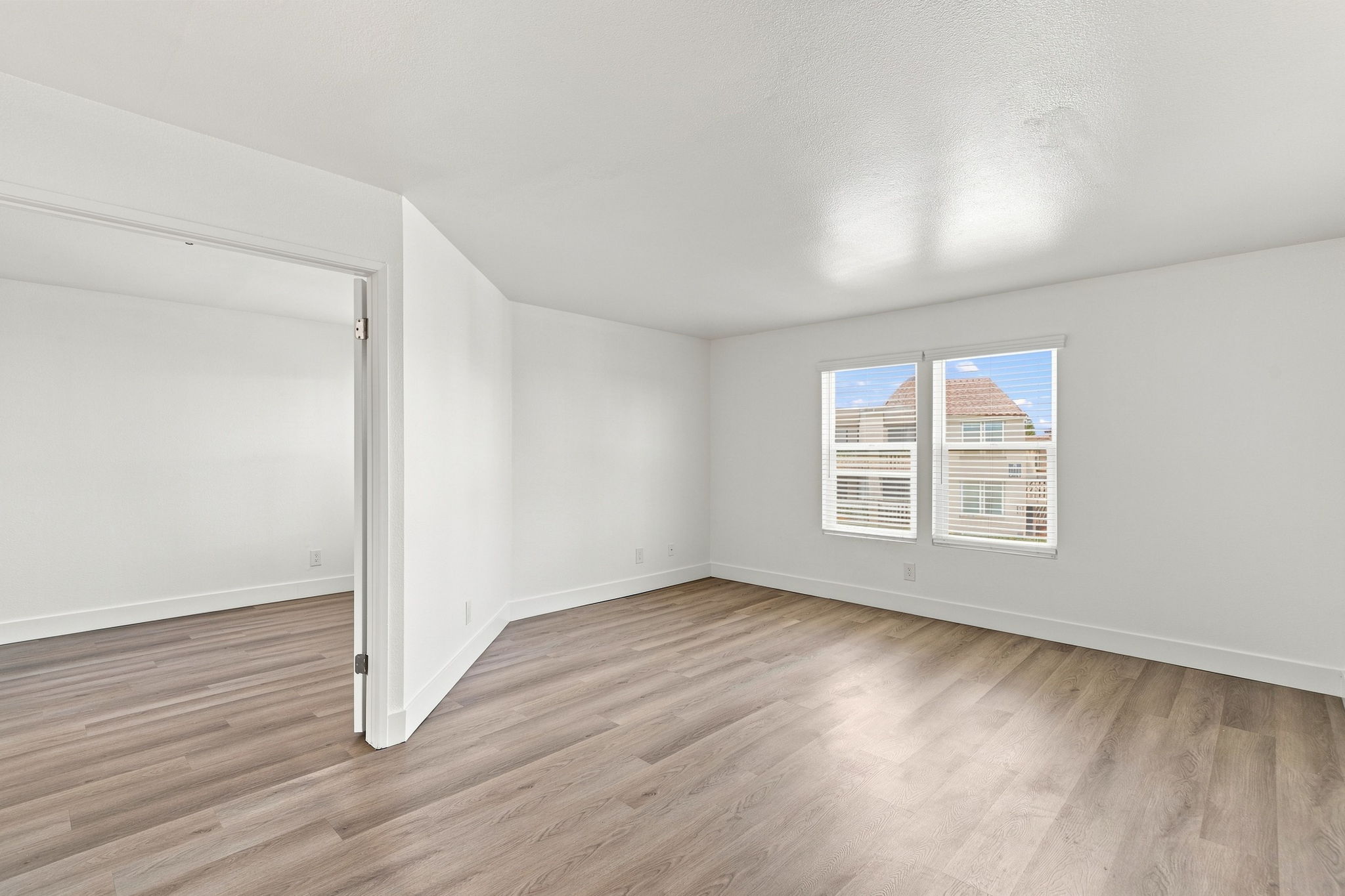 A bright and airy room with light-colored walls and wooden flooring. Two windows allow natural light to flood in, showcasing a peaceful view outside. The space is empty, suggesting it could be used for various purposes, such as a living area or office. A doorway leads to another similar room.
