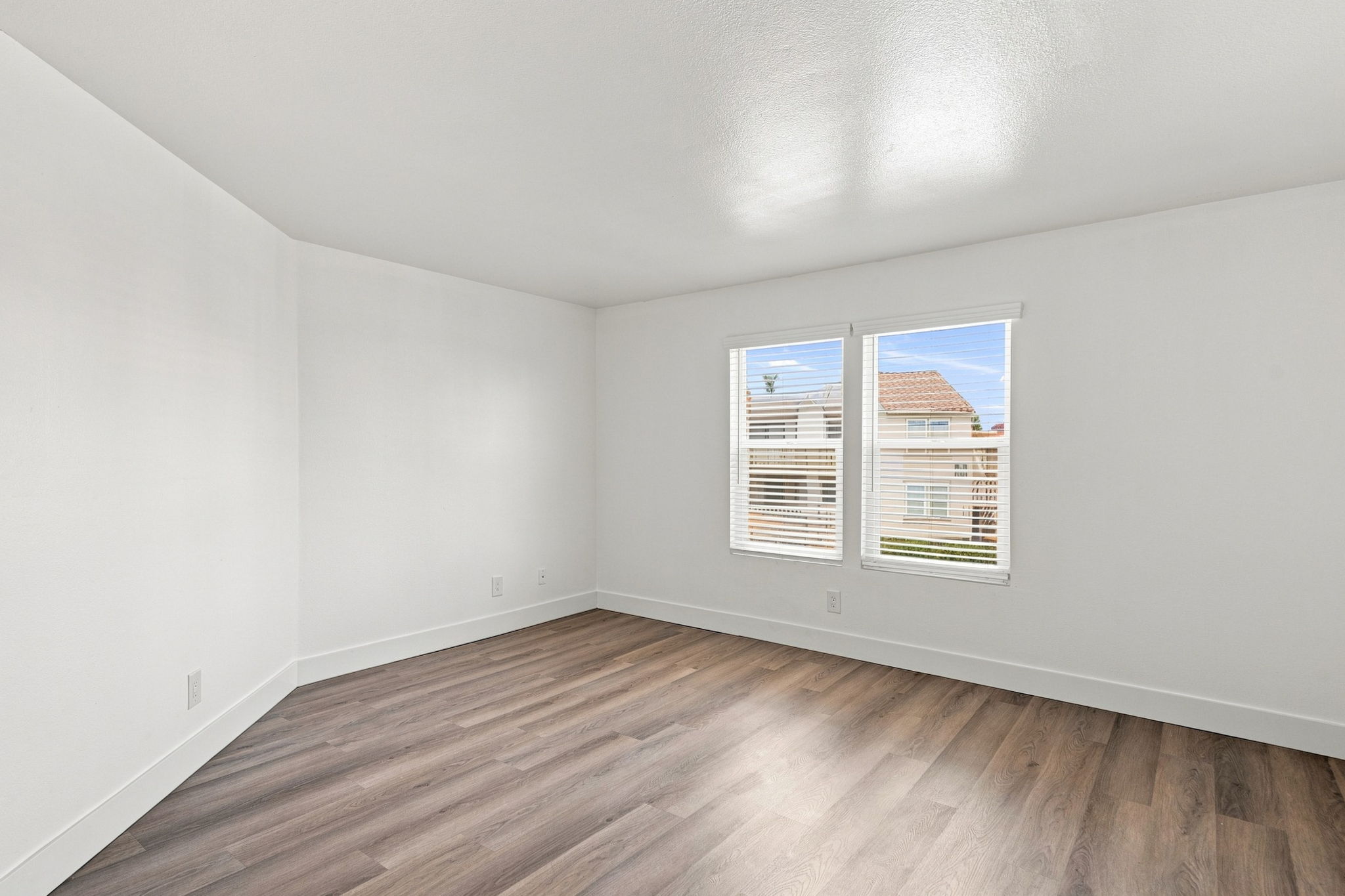 An empty room with white walls and a wooden floor. Two windows with blinds let in natural light, revealing a view of neighboring buildings outside. The room has no furnishings or decorations, creating an open and spacious feel.