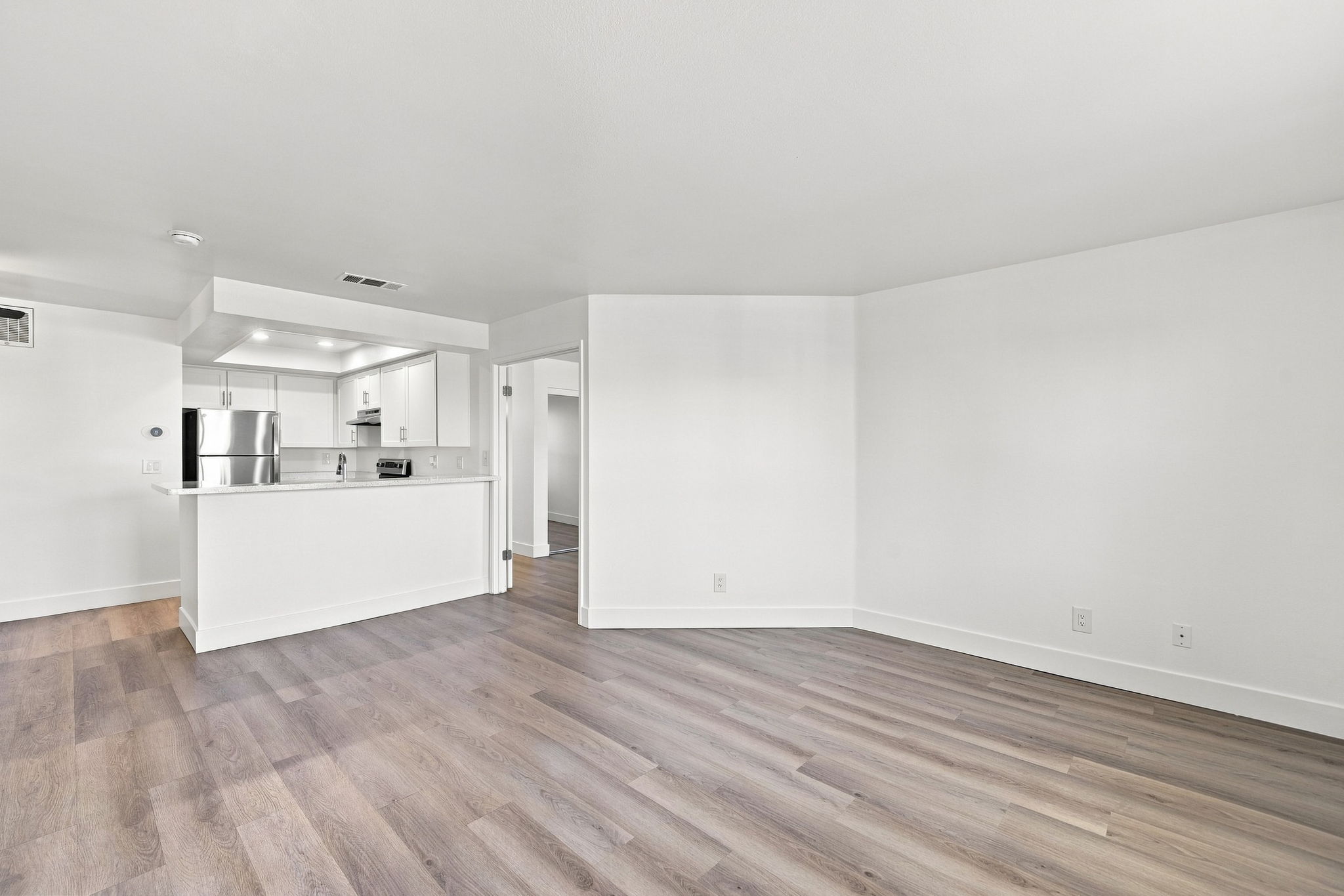 A modern, empty living space with light-colored walls and hardwood flooring. The room features a doorway leading to another area, a small kitchen visible in the background with white cabinetry and appliances, and plenty of natural light.