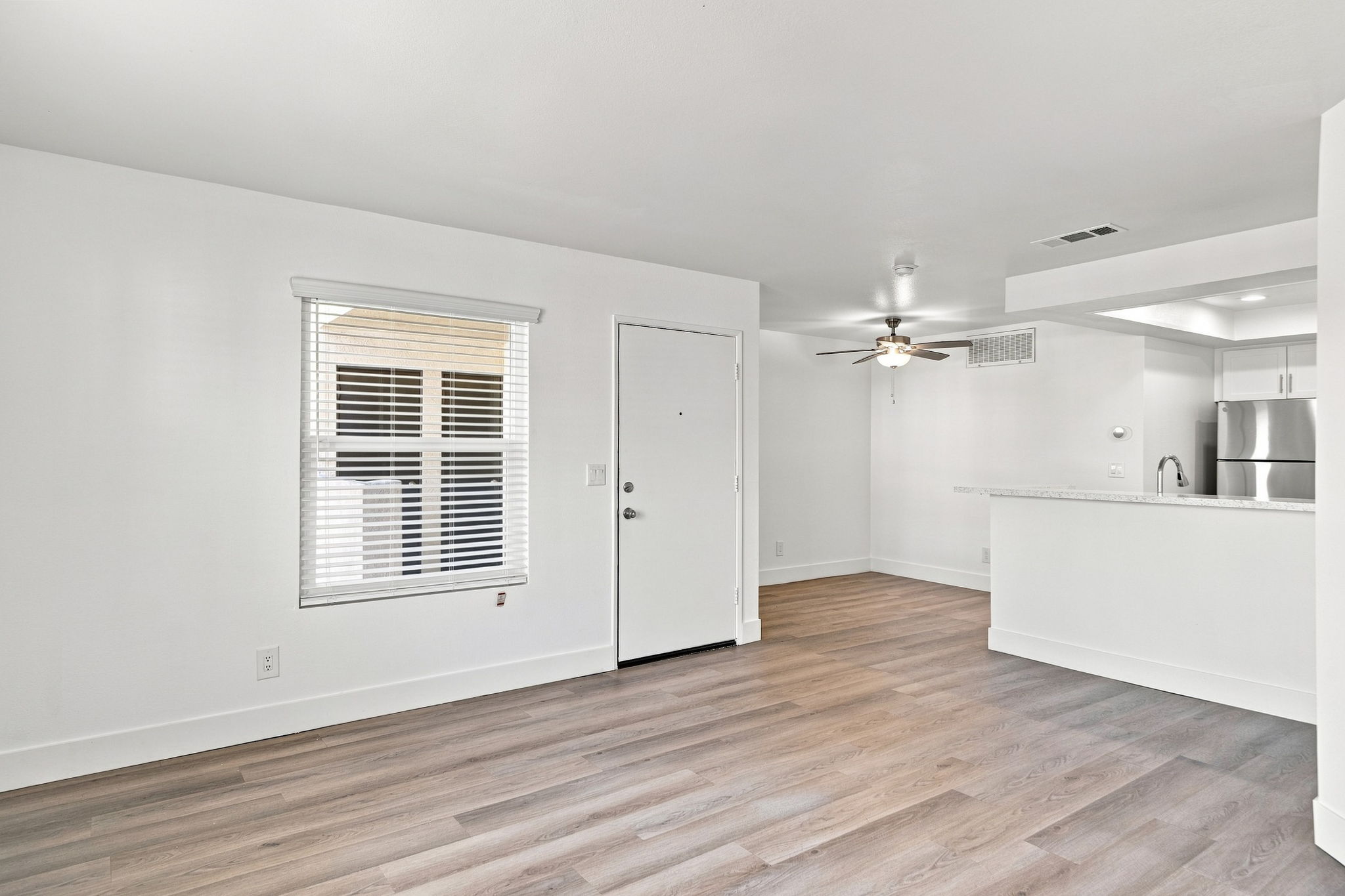 A bright and modern living space with light-colored walls, wooden flooring, and a ceiling fan. A window with white blinds allows natural light in. To the right, there's an open kitchen area with a counter. The entrance door is visible in the foreground. The overall atmosphere is clean and inviting.