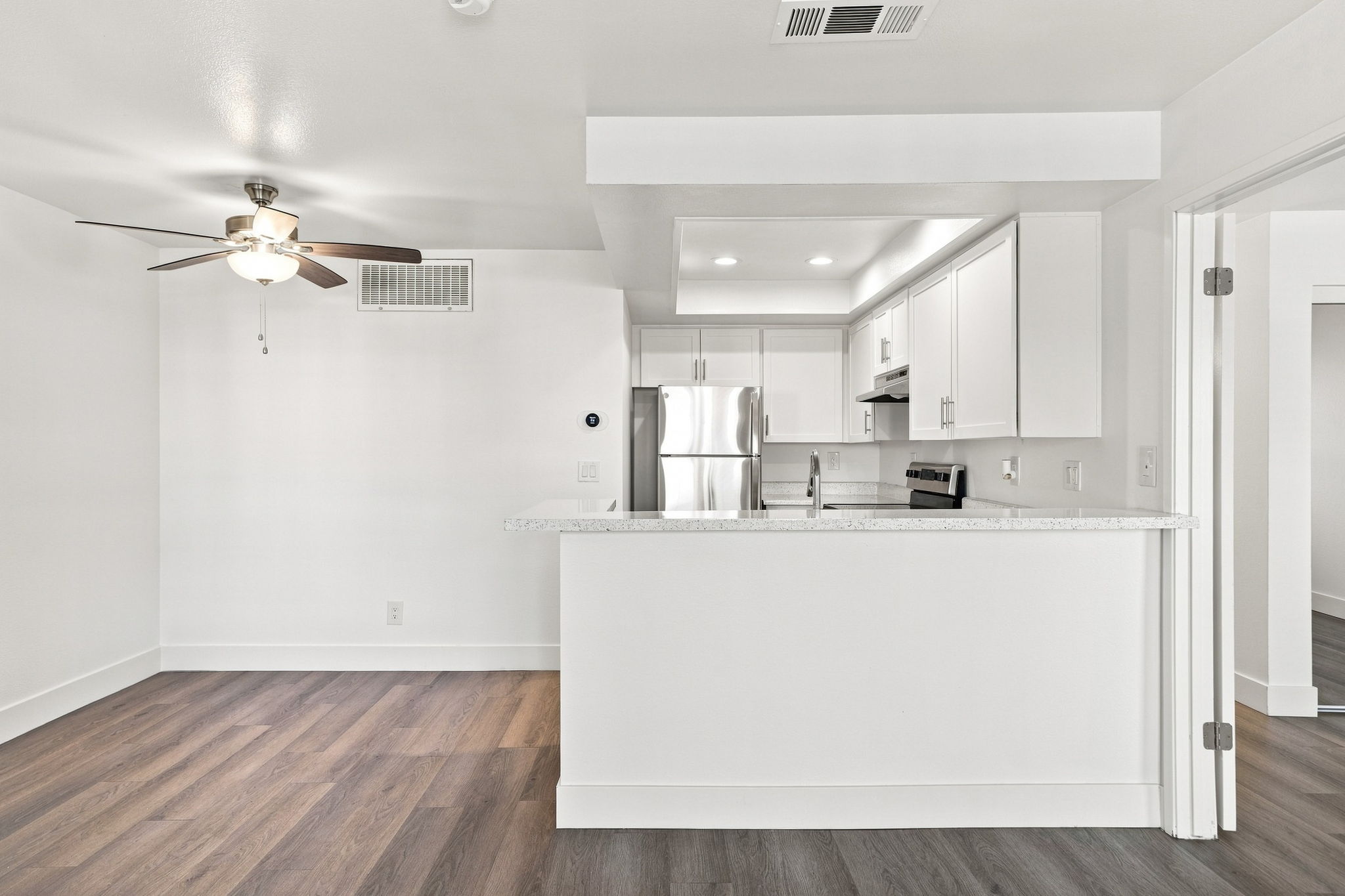 Interior of a modern, bright kitchen and living area. Features a ceiling fan, stainless steel appliances, and white cabinetry. The space has a clean, open layout with warm wood-style flooring and is well-lit, creating an inviting atmosphere.