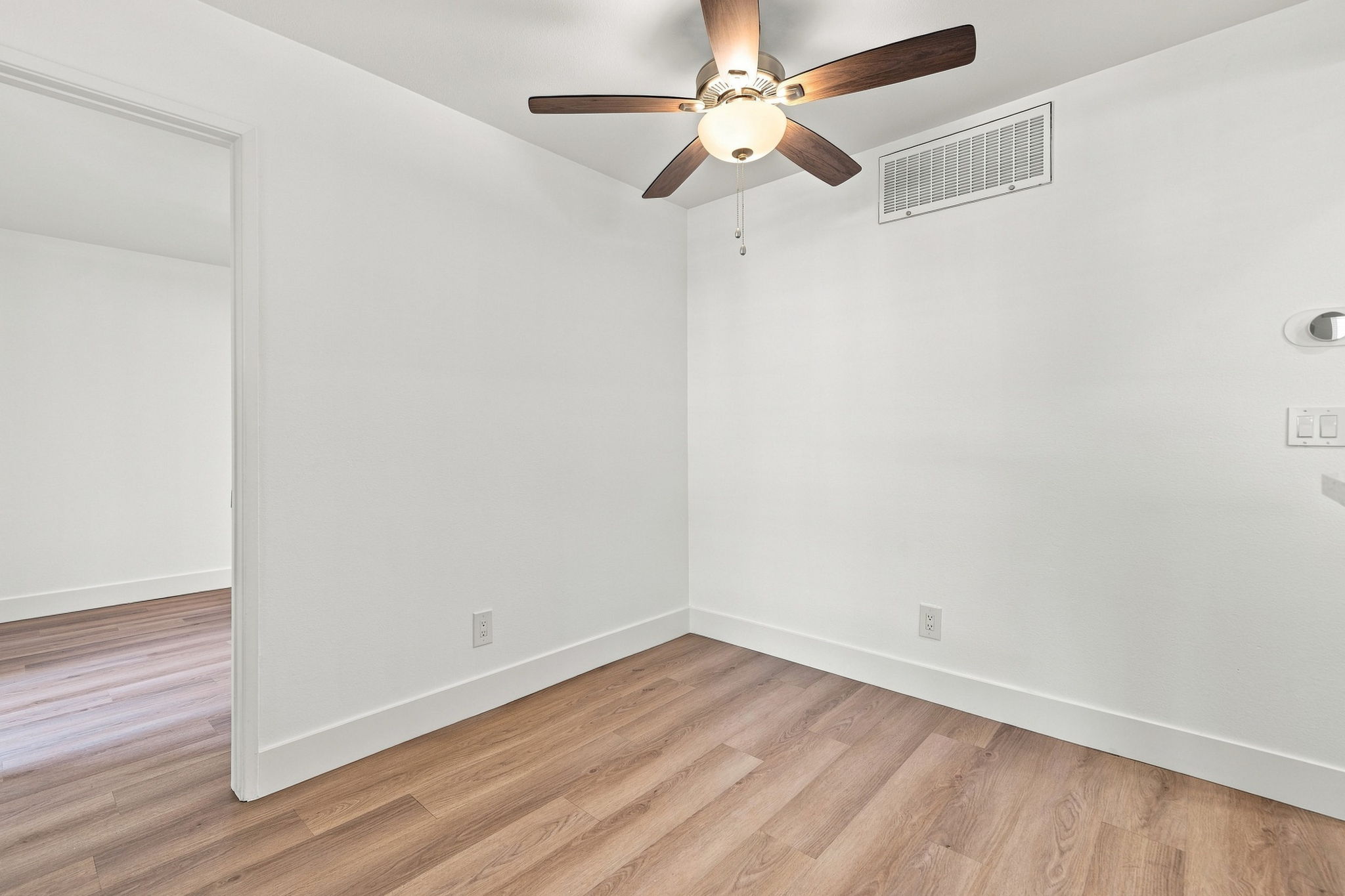 Interior view of a clean, empty room with light-colored walls and hardwood flooring. A ceiling fan with wooden blades hangs from the ceiling. An air vent is visible on the wall. A doorway leads to another room, suggesting a spacious layout. Natural light enhances the open feel of the space.