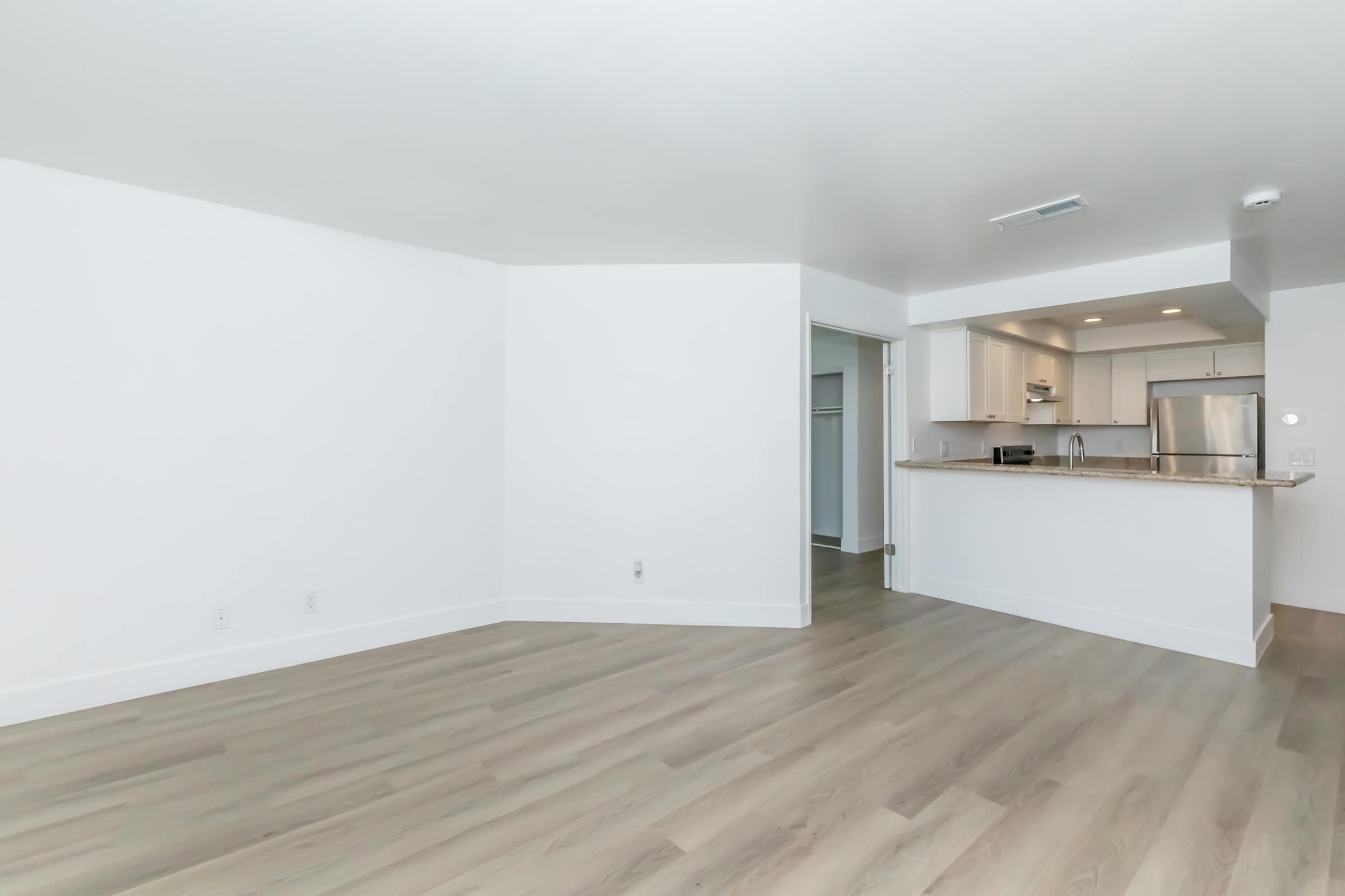 A spacious, modern living area with light-colored hardwood flooring and white walls. The room features a doorway leading to a kitchen with stainless steel appliances and a countertop. Natural light enhances the bright and airy atmosphere, making it feel open and inviting.