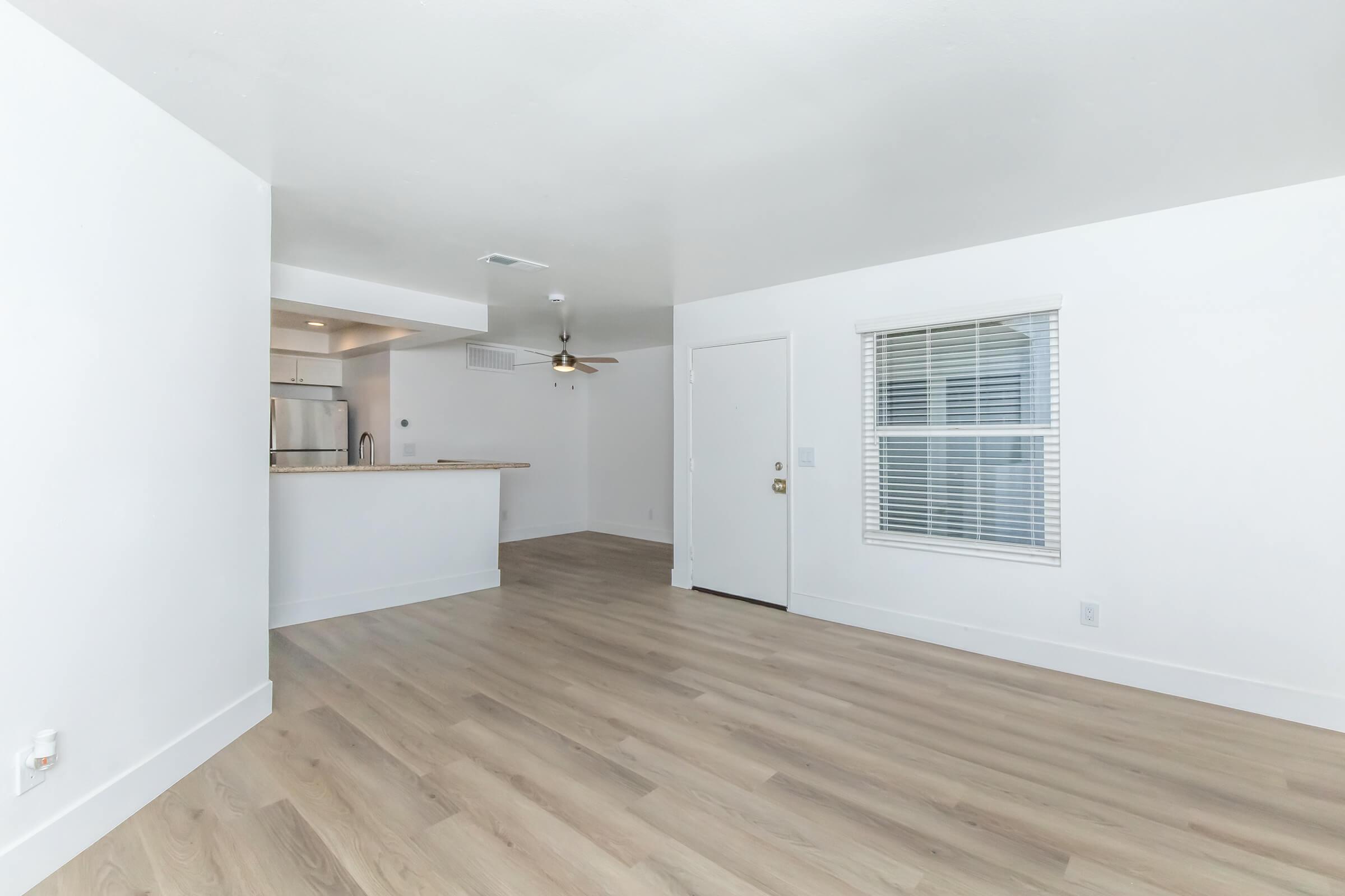 A light and airy living space featuring smooth wooden flooring, white walls, and large windows letting in natural light. A door leads outside, and a kitchen area with a counter is visible in the background, complemented by modern appliances and a ceiling fan.