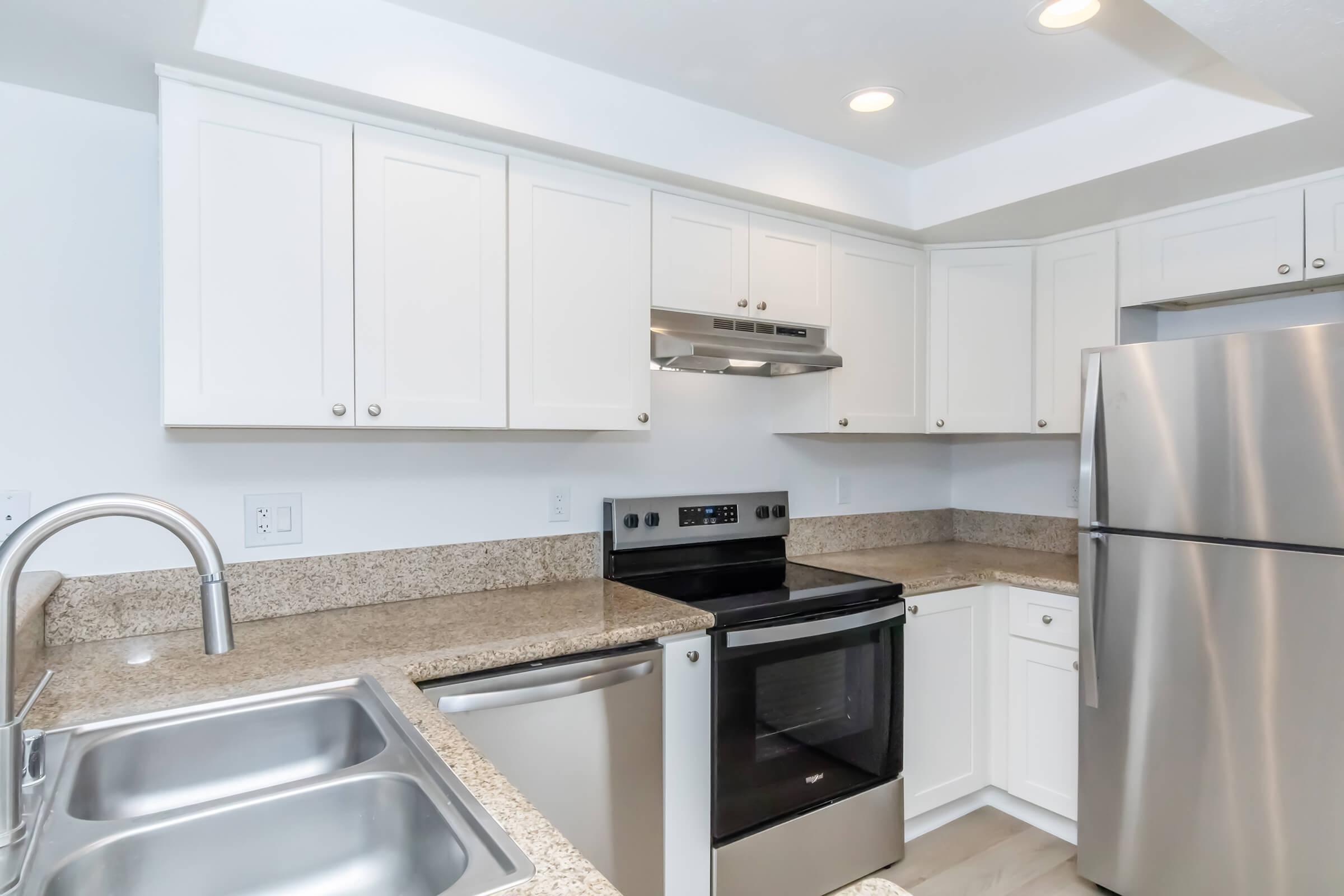 Modern kitchen featuring white cabinets, stainless steel appliances, and a granite countertop. Includes a double sink and a glass-top stove, with an oven beneath. The compact design offers an efficient use of space, with a refrigerator and an exhaust hood above the stove. Bright overhead lighting enhances the atmosphere.