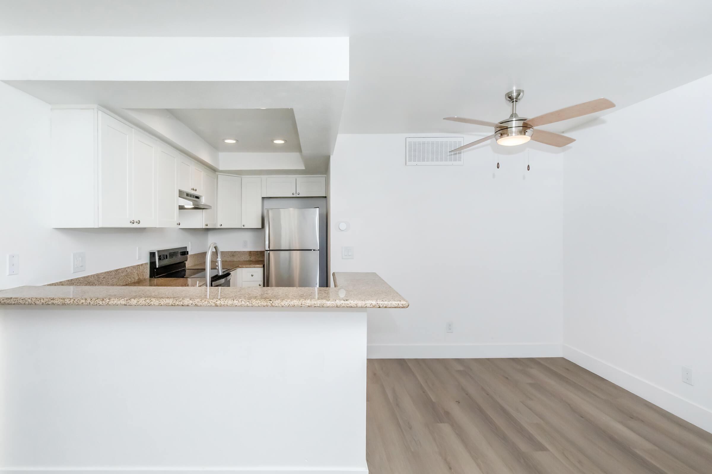 Modern kitchen and living area featuring a granite countertop breakfast bar, stainless steel appliances, a ceiling fan, and light wood flooring. The walls are painted white, creating a bright and spacious atmosphere.
