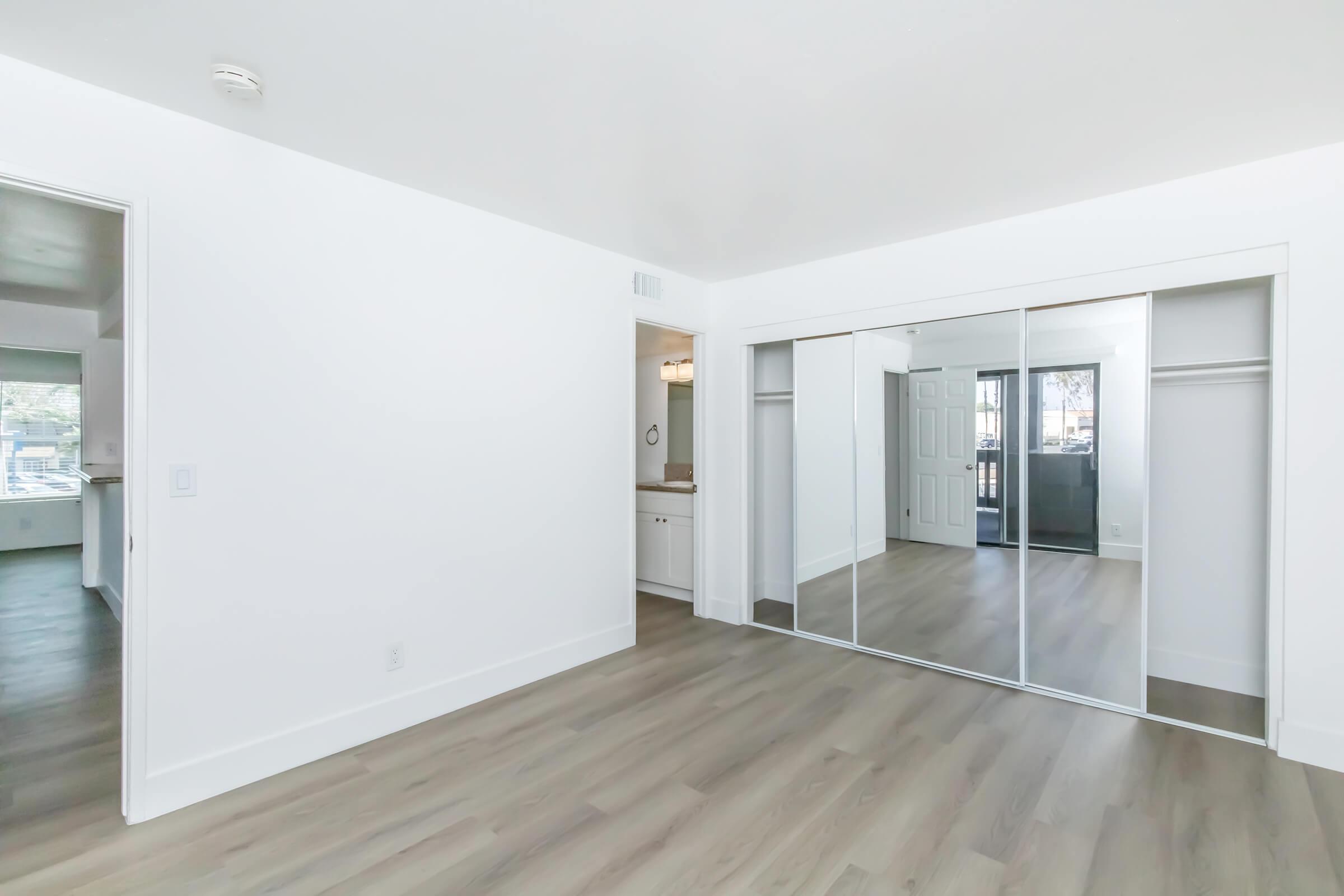 Bright, modern interior of a room featuring light wood flooring, mirrored closet doors, and white walls. The space includes an entryway leading to a kitchen area visible in the background, with natural light coming through large windows. Overall, the room appears minimalist and well-maintained.