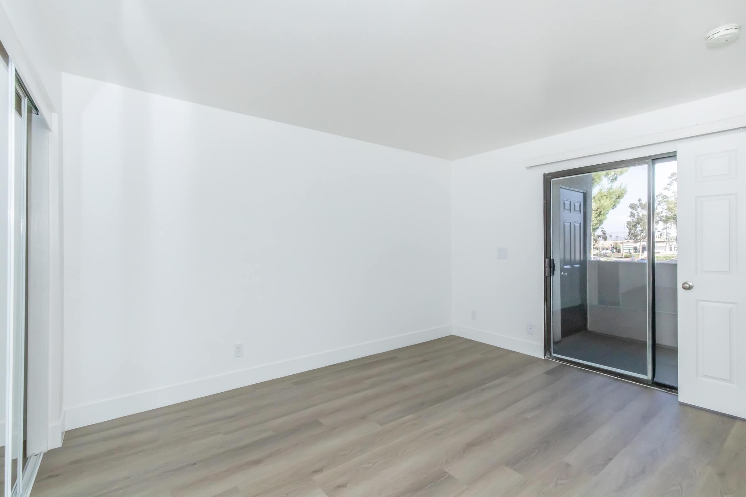 Empty interior of a room with white walls and light wood flooring. A large sliding glass door leads to a balcony outside. Natural light streams in from the door, creating a bright and open atmosphere. No furniture or decorations are present in the space.