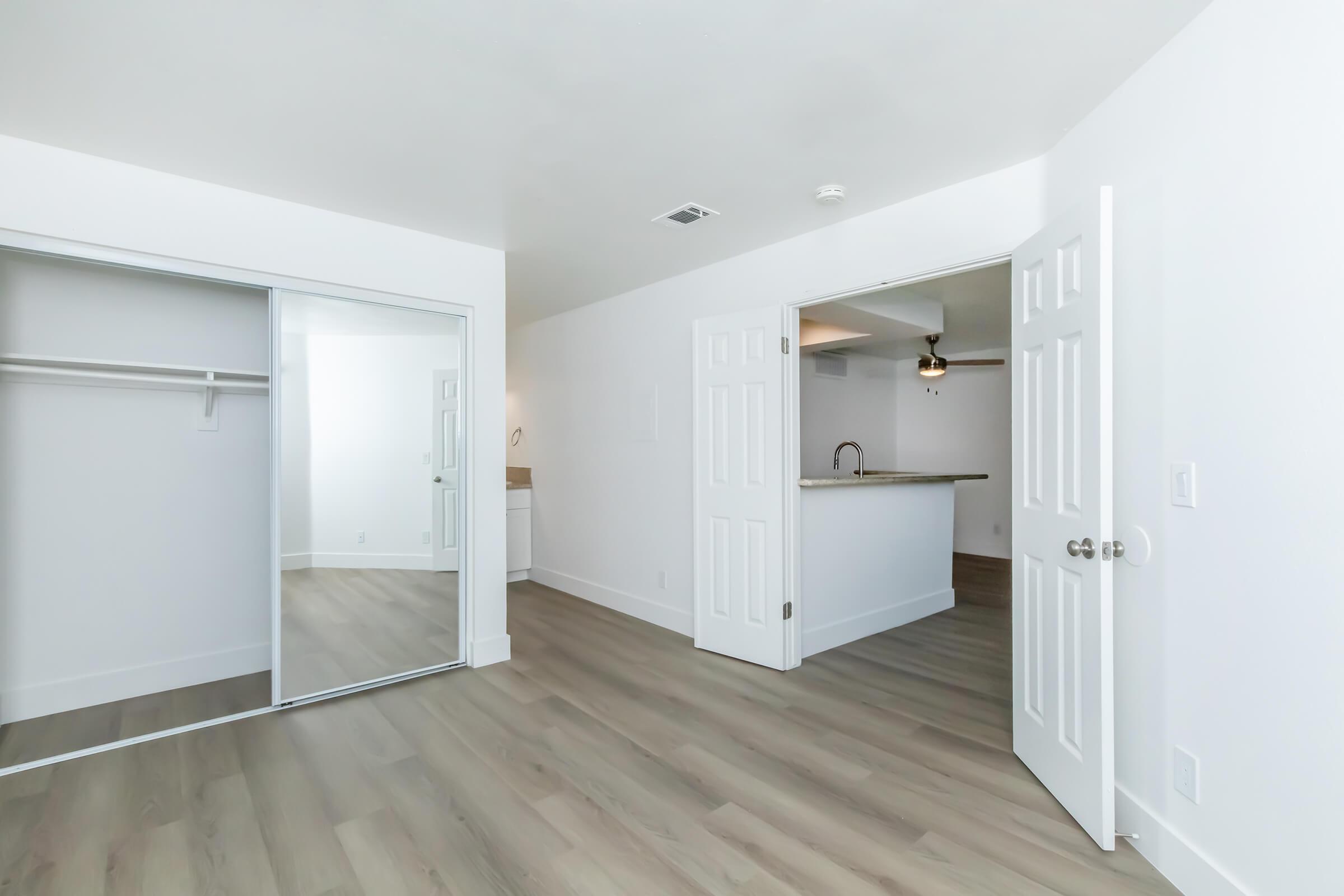 A spacious, well-lit room with light wood flooring. There is a mirrored closet on the left and an open doorway leading to a kitchen area. The walls are painted white, and natural light brightens the space. The overall design is modern and minimalistic.