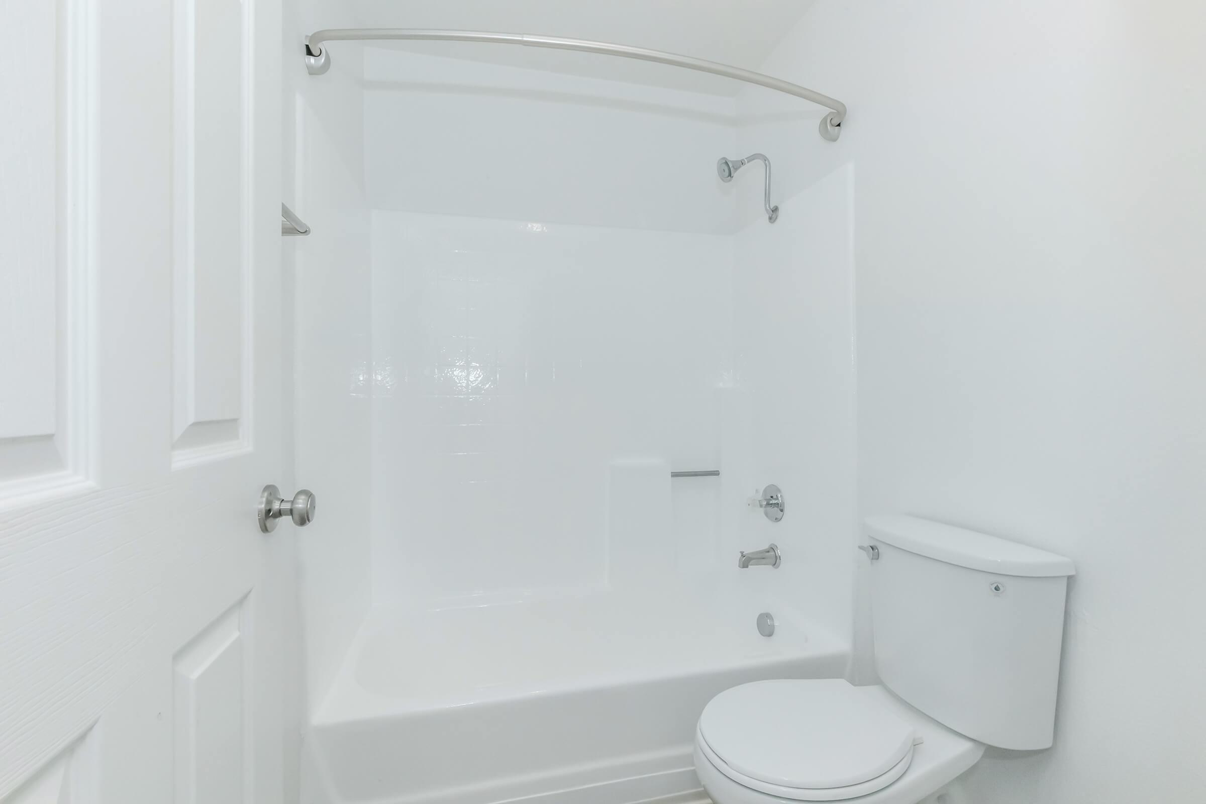 A clean and modern bathroom featuring a white bathtub with a showerhead, a white toilet, and a sliding door. The walls are painted white, creating a bright and spacious feel. The overall design is simple and functional.