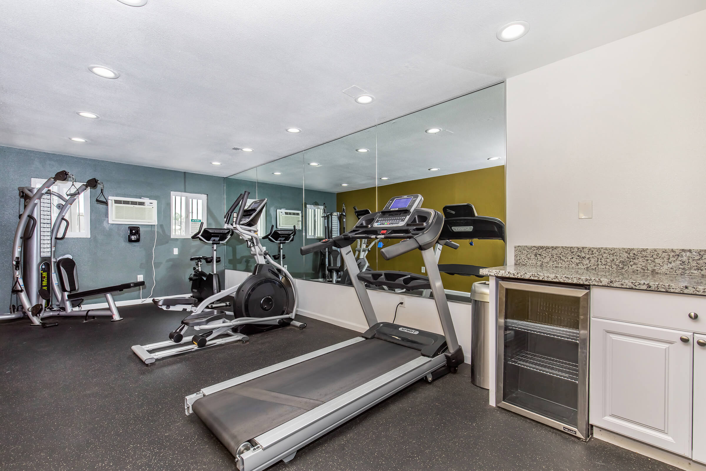 A modern gym interior featuring a treadmill, elliptical machine, and a cable machine. The walls are painted in two colors, and large mirrors reflect the gym equipment. There's also a small fridge and countertop area on one side, with overhead lighting providing a bright atmosphere.