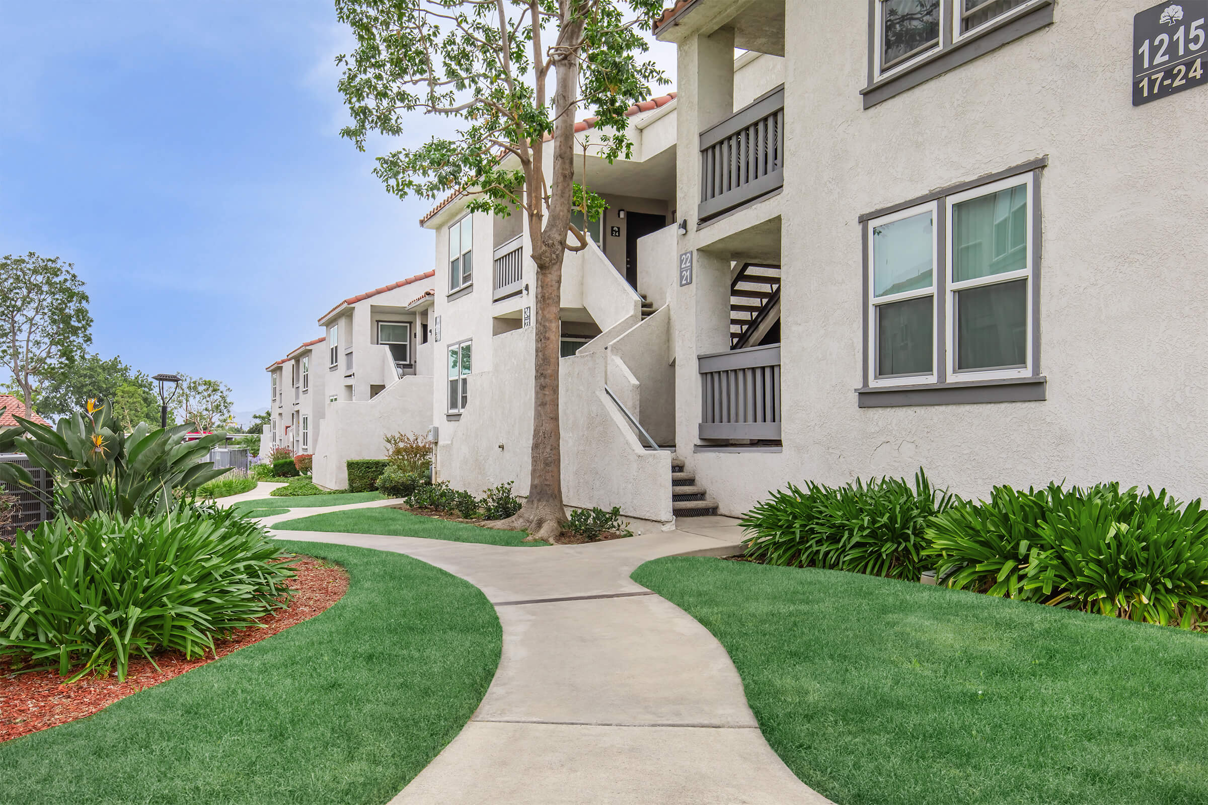 A neatly landscaped pathway leading through a residential complex, featuring white apartment buildings with gray trim. Lush green grass and colorful plants line the walkway, while a tree adds shade. The scene is bright and inviting, showcasing a well-maintained outdoor area.