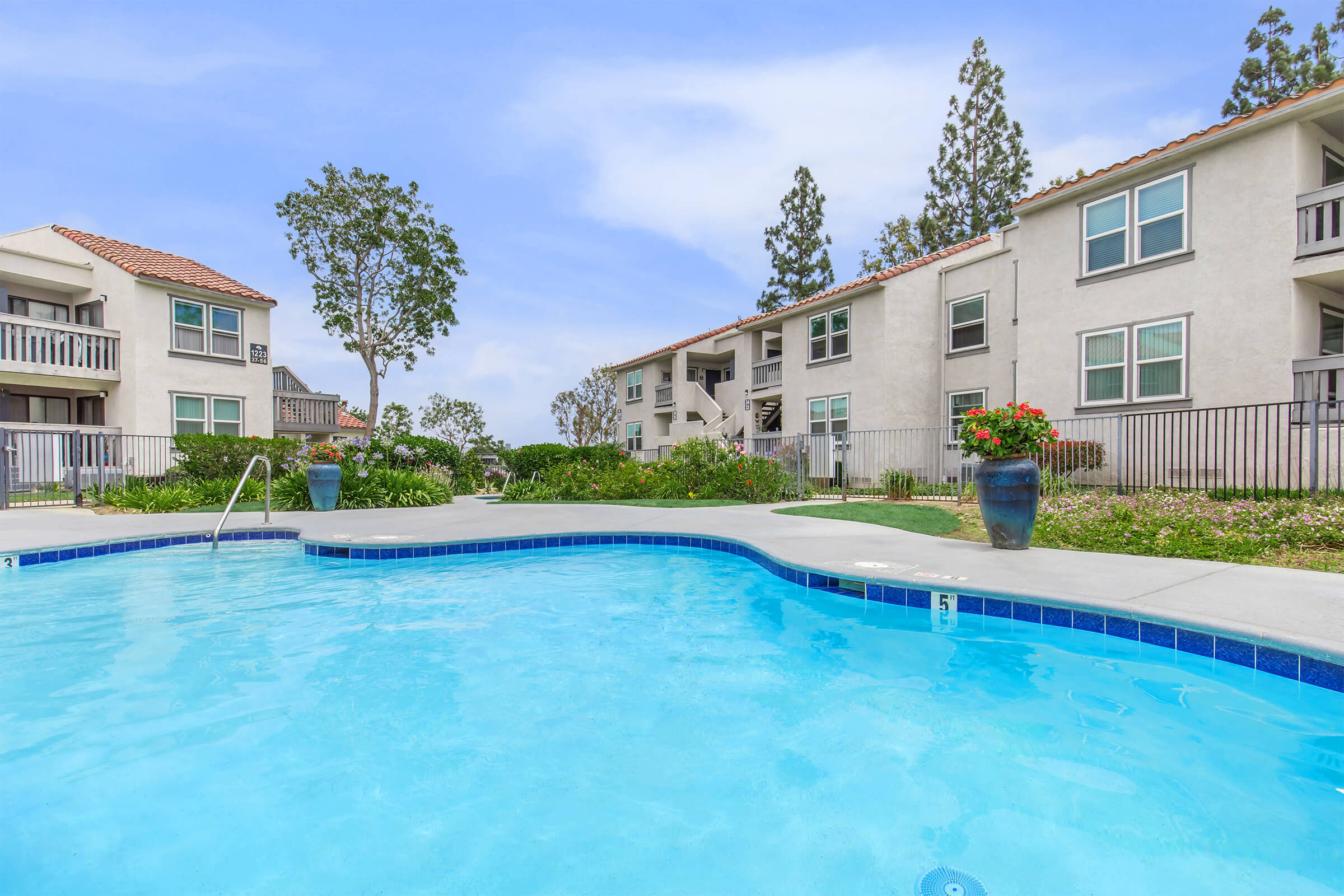 A sparkling blue swimming pool surrounded by well-maintained greenery, flowers, and several apartment buildings in the background. The scene is bright and welcoming, evoking a refreshing outdoor atmosphere. The pool area is equipped with steps leading into the water and decorative planters.