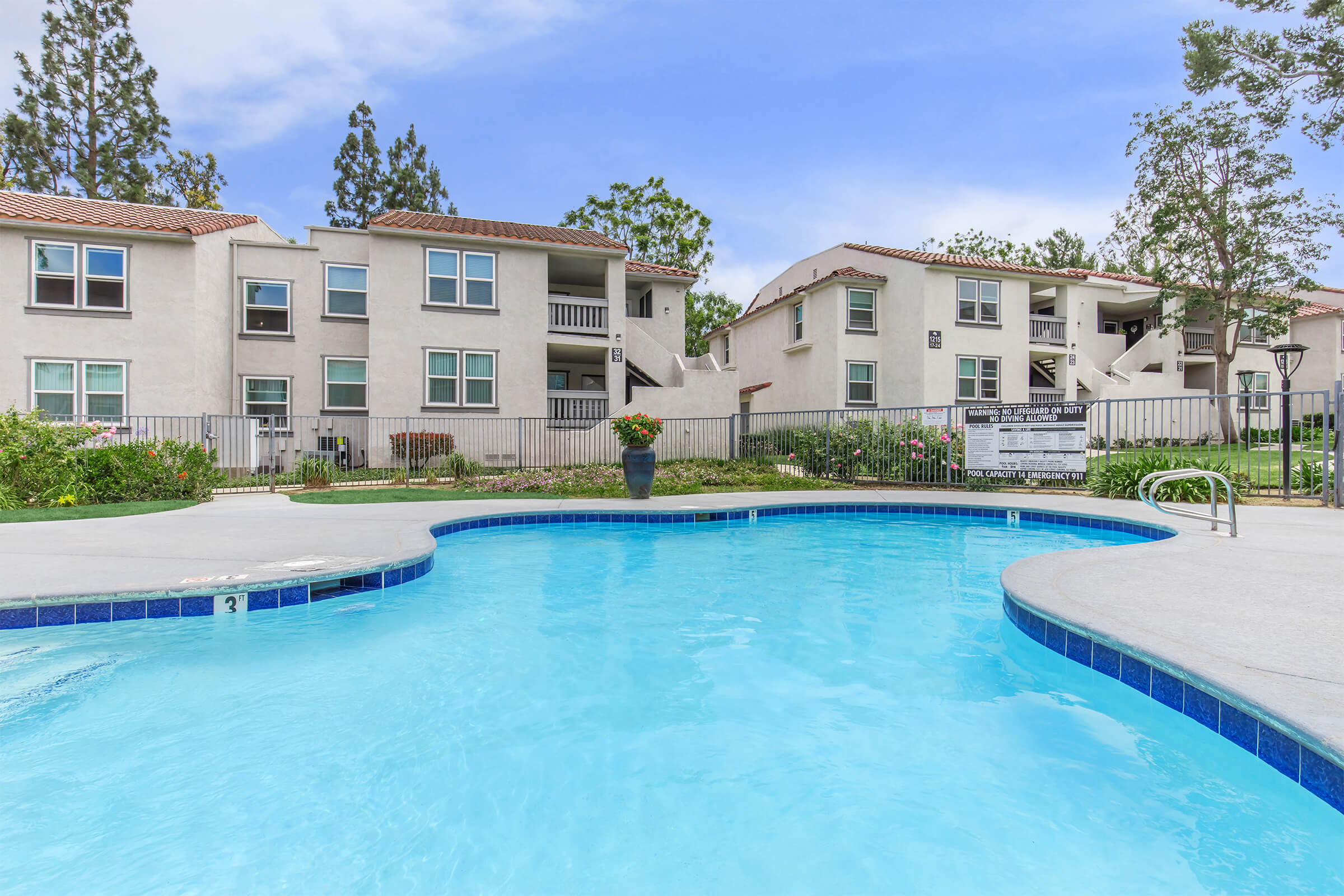 A clear blue swimming pool in the foreground with steps leading in, surrounded by a landscaped area. In the background, there are two-story apartment buildings with balconies and green trees. The sky is bright and partly cloudy, creating a welcoming outdoor atmosphere.