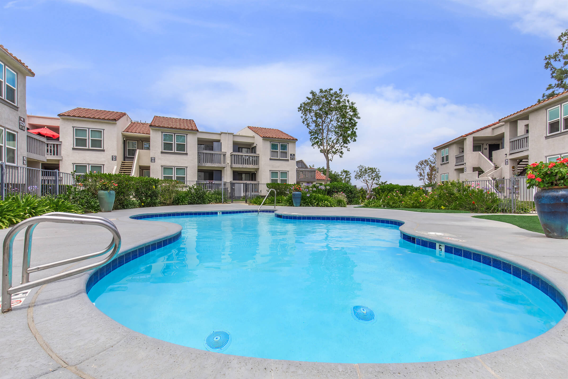 A clear swimming pool shaped like a kidney, surrounded by a landscaped area with green plants and trees. In the background, there are multi-story apartment buildings with balconies and a blue sky overhead. Red umbrellas are set up near the pool area, creating a relaxed atmosphere.