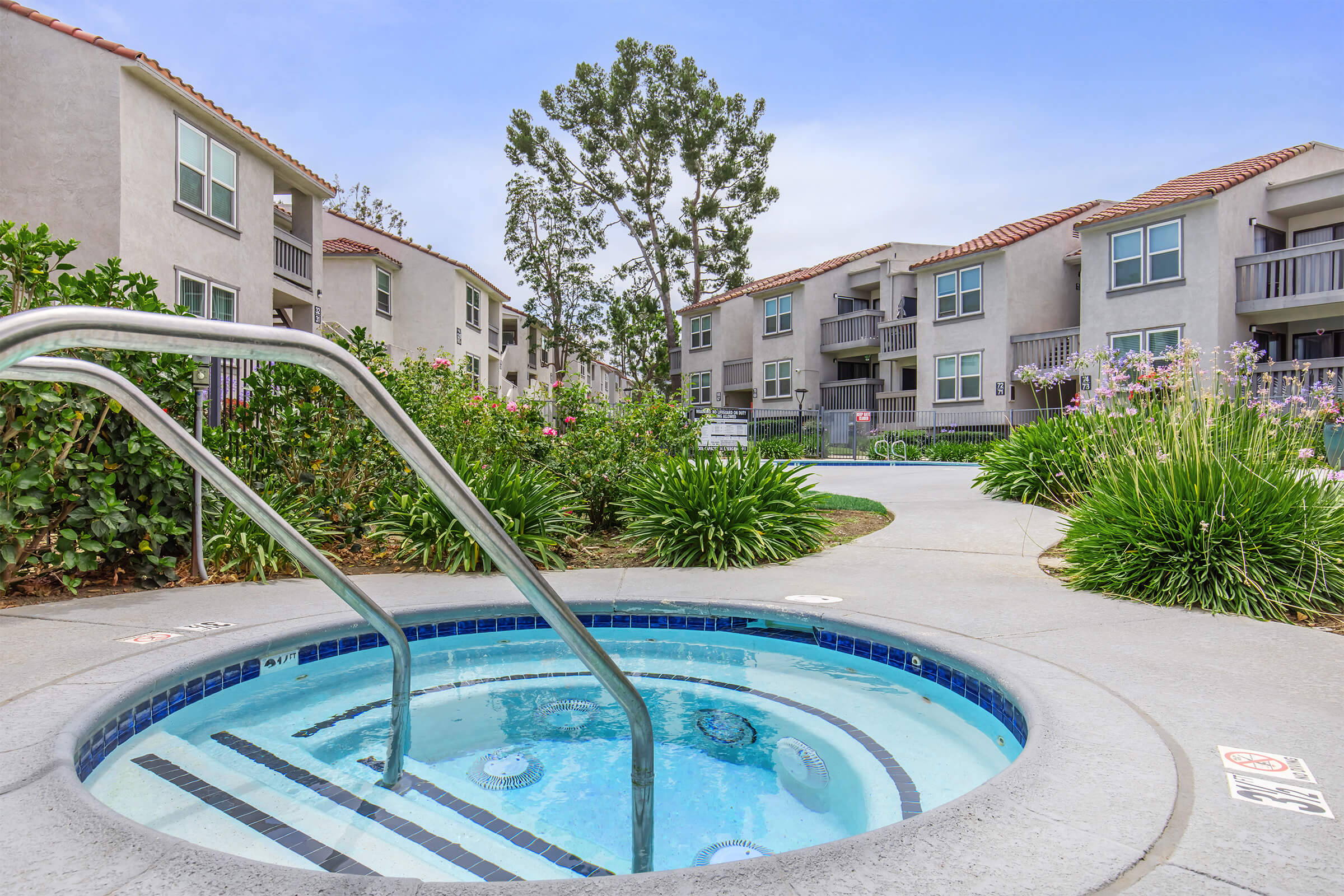 A view of a jacuzzi with steps leading in, surrounded by lush green landscaping. In the background, several apartment buildings are visible with balconies and multiple windows. The sky is clear, indicating a sunny day.
