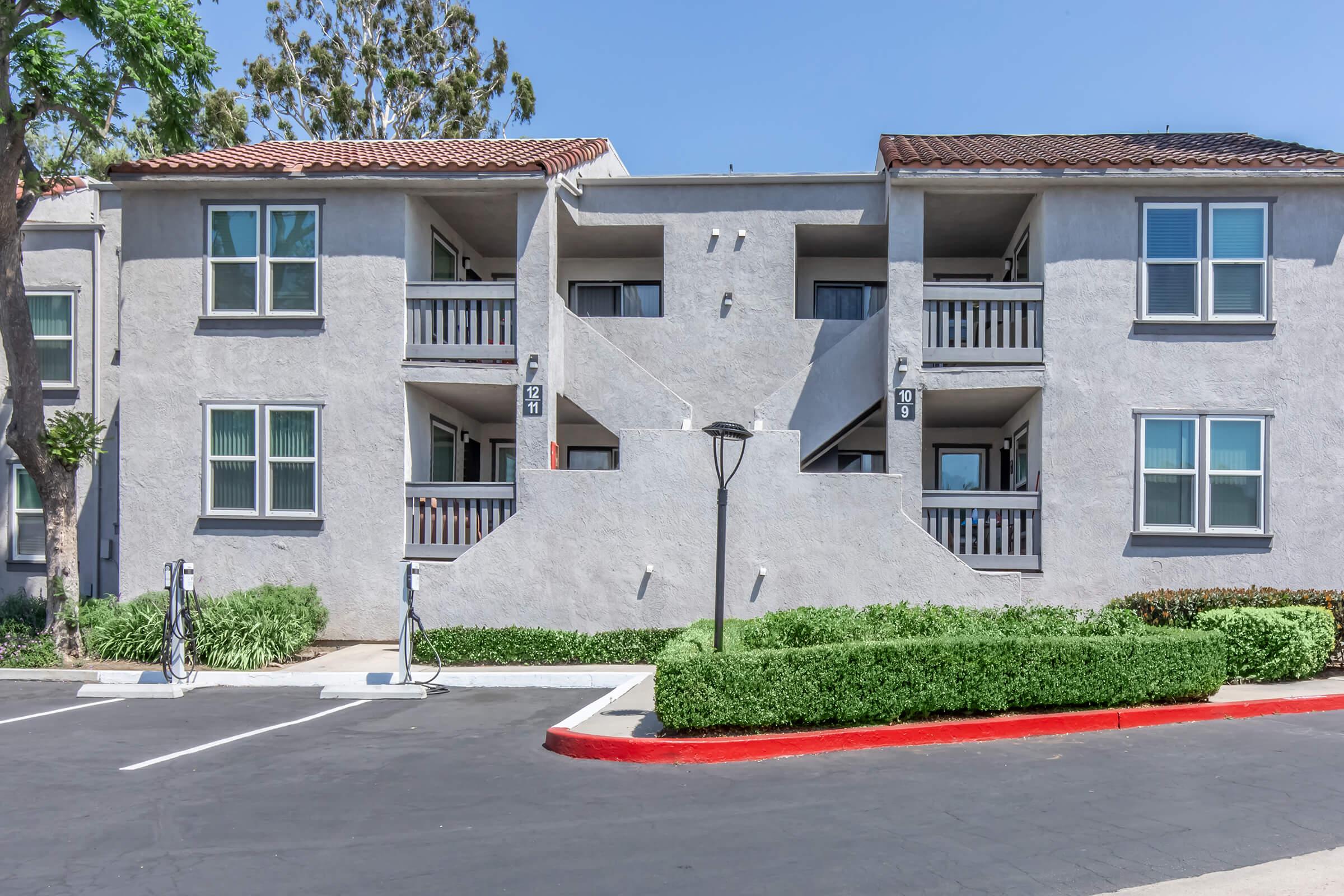 A multi-unit residential building featuring a gray exterior. The structure has multiple balconies and windows, with a landscaped area and parking spaces visible in the foreground. The design includes a red curb and a lamppost, surrounded by greenery. The sky is clear and blue.