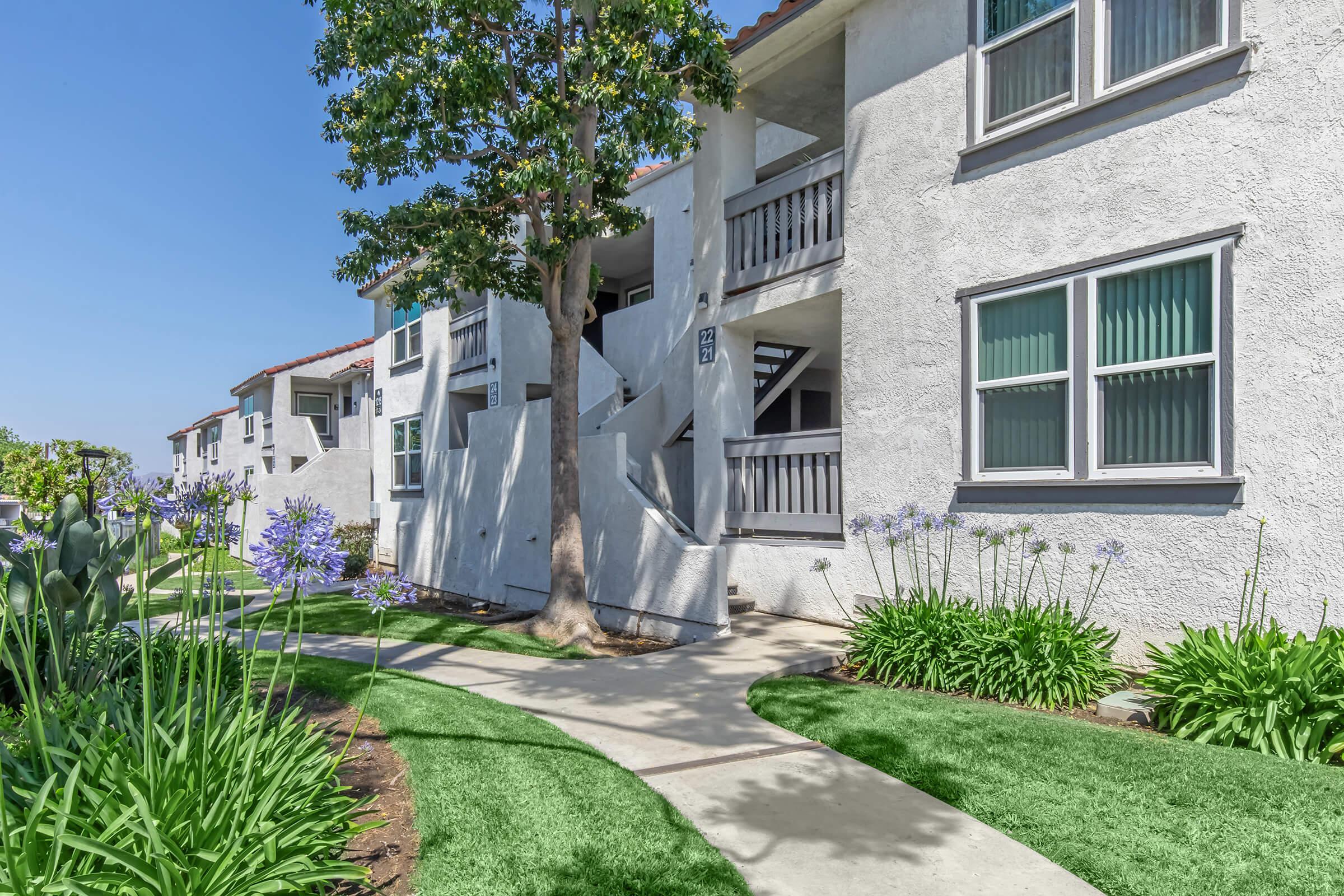 A sunny view of an apartment complex featuring white stucco buildings with balconies, surrounded by green landscaping. A paved pathway winds through the garden, lined with purple flowers and lush grass. Trees provide shade, creating a welcoming outdoor space.