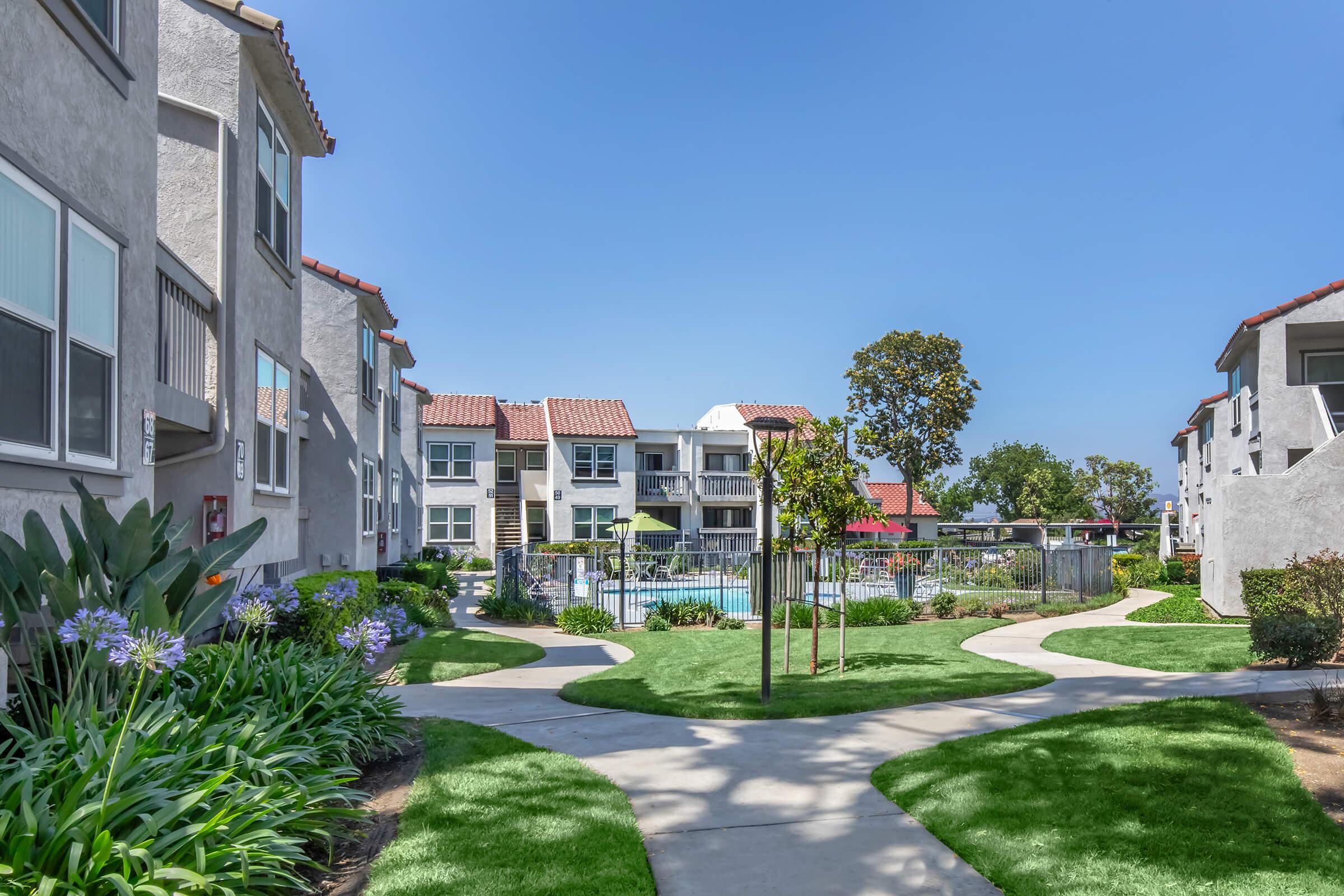 A landscaped pathway in an apartment complex leads to a swimming pool area. The scene features well-maintained grass, trees, and flowering plants. Surrounding buildings have a Mediterranean architectural style with red-tiled roofs, creating a peaceful and inviting outdoor environment.