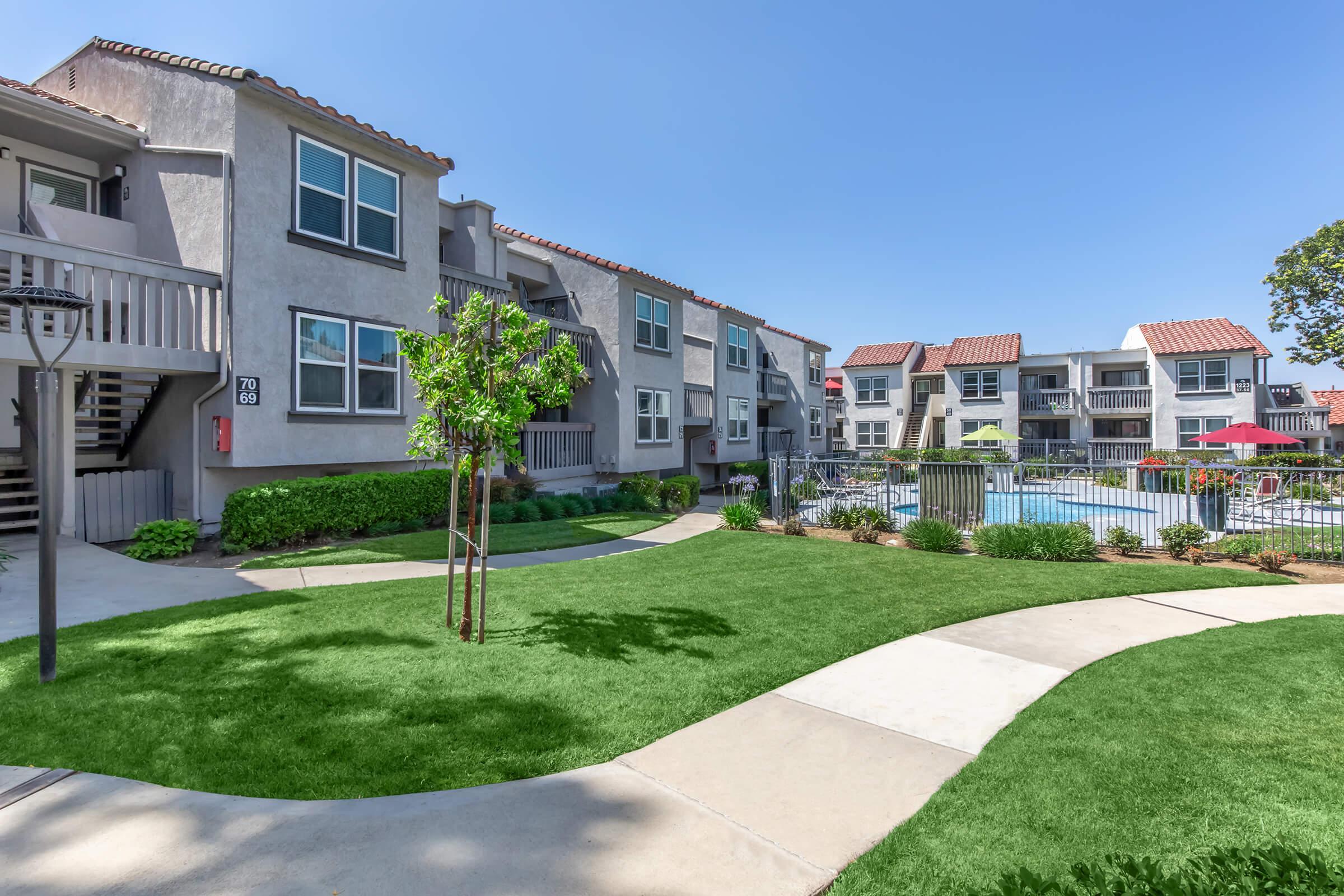 A residential apartment complex featuring two-story buildings with balconies, surrounded by well-maintained lawns and walking paths. In the background, there's a swimming pool area with lounge chairs and umbrellas, set against a clear blue sky.