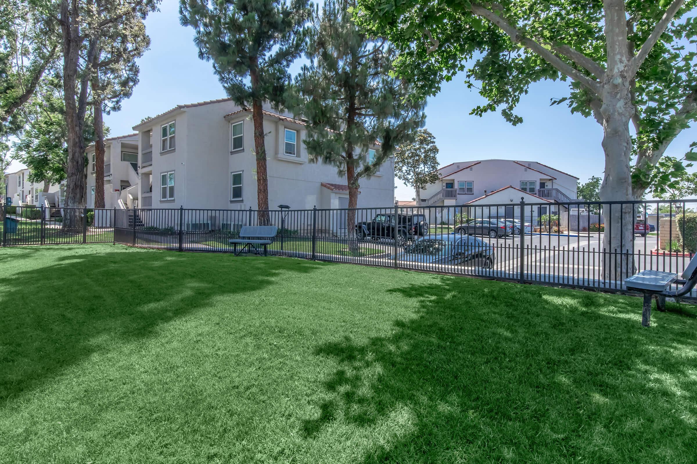A green lawn area with a black fence in the foreground. Behind the fence, there are two residential buildings surrounded by trees, with a parking area visible. The scene is sunny, highlighting the greenery and the well-kept surroundings.
