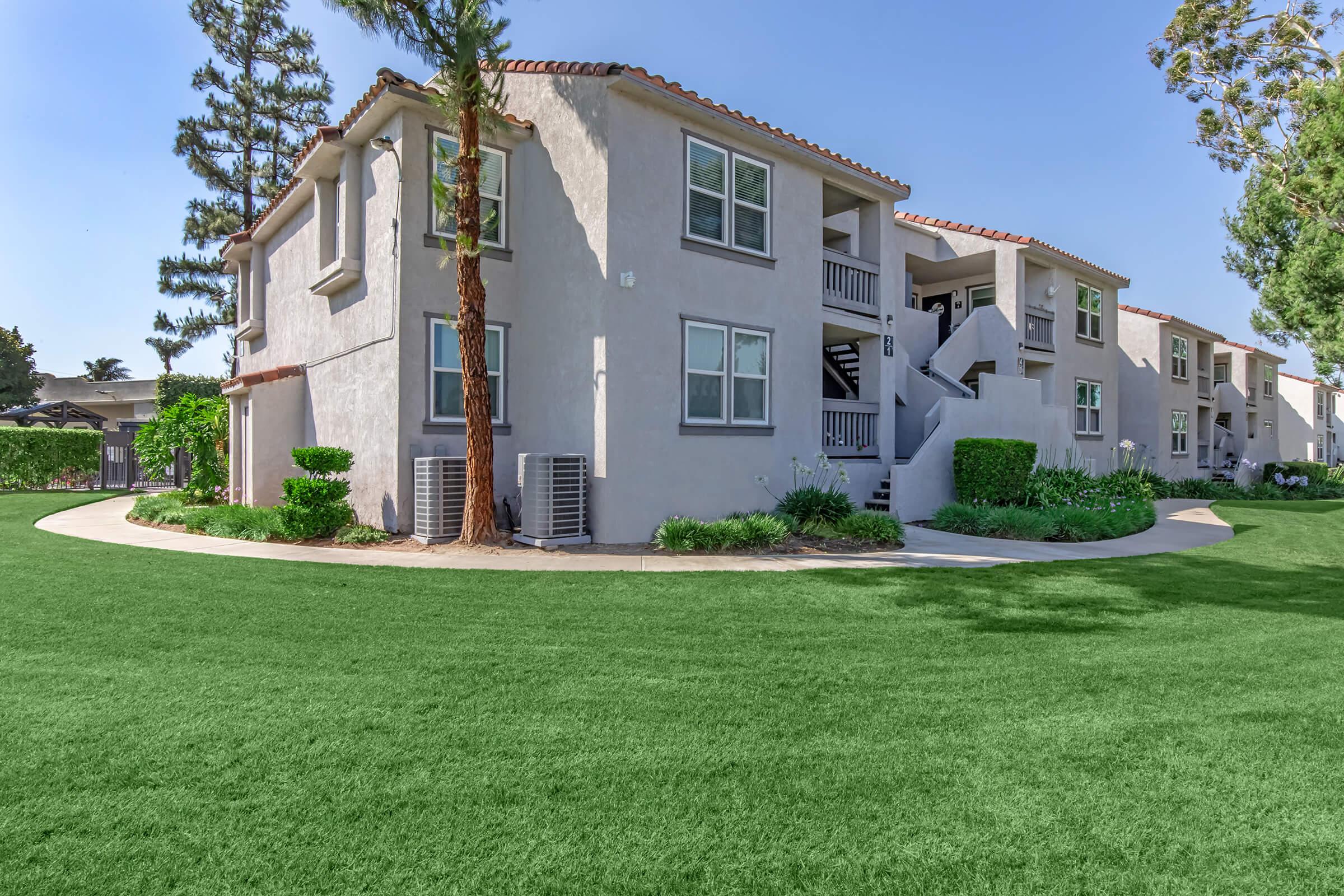 A well-maintained multi-unit residential building with a light gray exterior, surrounded by greenery and manicured lawns. There are air conditioning units visible, and the landscape includes palm trees and a curved walking path. The sky is clear and blue, enhancing the inviting atmosphere of the property.