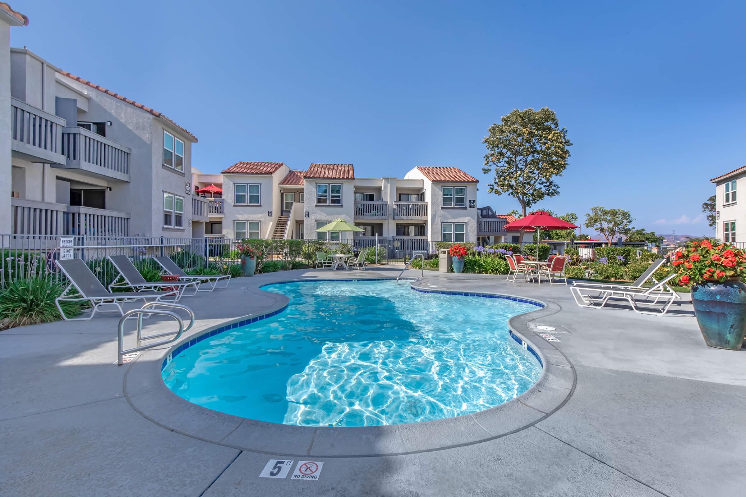 A brightly lit outdoor swimming pool surrounded by lounge chairs and potted plants. Nearby, there are colorful umbrellas providing shade, and apartments with balconies are visible in the background under a clear blue sky. The scene conveys a relaxing atmosphere ideal for leisure activities.