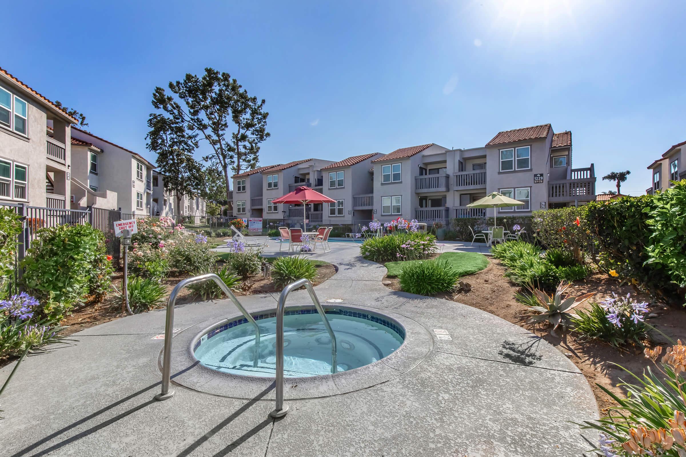 A vibrant outdoor space featuring a hot tub, surrounded by colorful flowers and shrubs. In the background, there are residential buildings with balconies, and a pool area with lounge chairs and umbrellas, set against a clear blue sky. The scene conveys a relaxing atmosphere ideal for leisure.