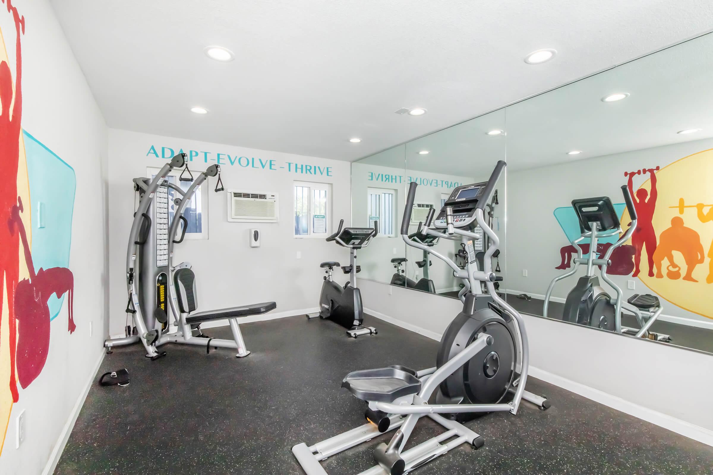 A small gym room featuring various exercise equipment, including a weight bench, elliptical machine, and a cable machine. The walls are decorated with motivational phrases, and large mirrors create an open feel. Bright, colorful graphics of people exercising add vibrancy to the space.