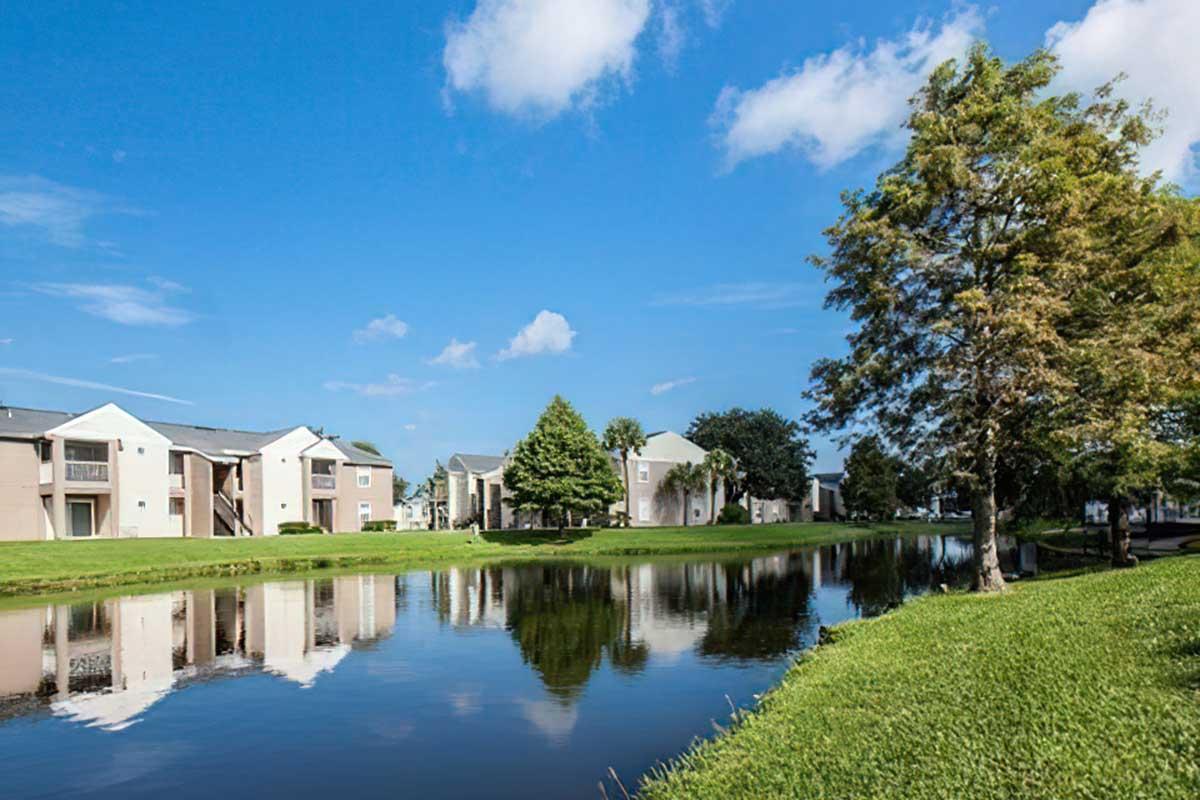 View of a peaceful residential area by a calm pond, featuring multiple two-story apartment buildings surrounded by lush greenery and trees.