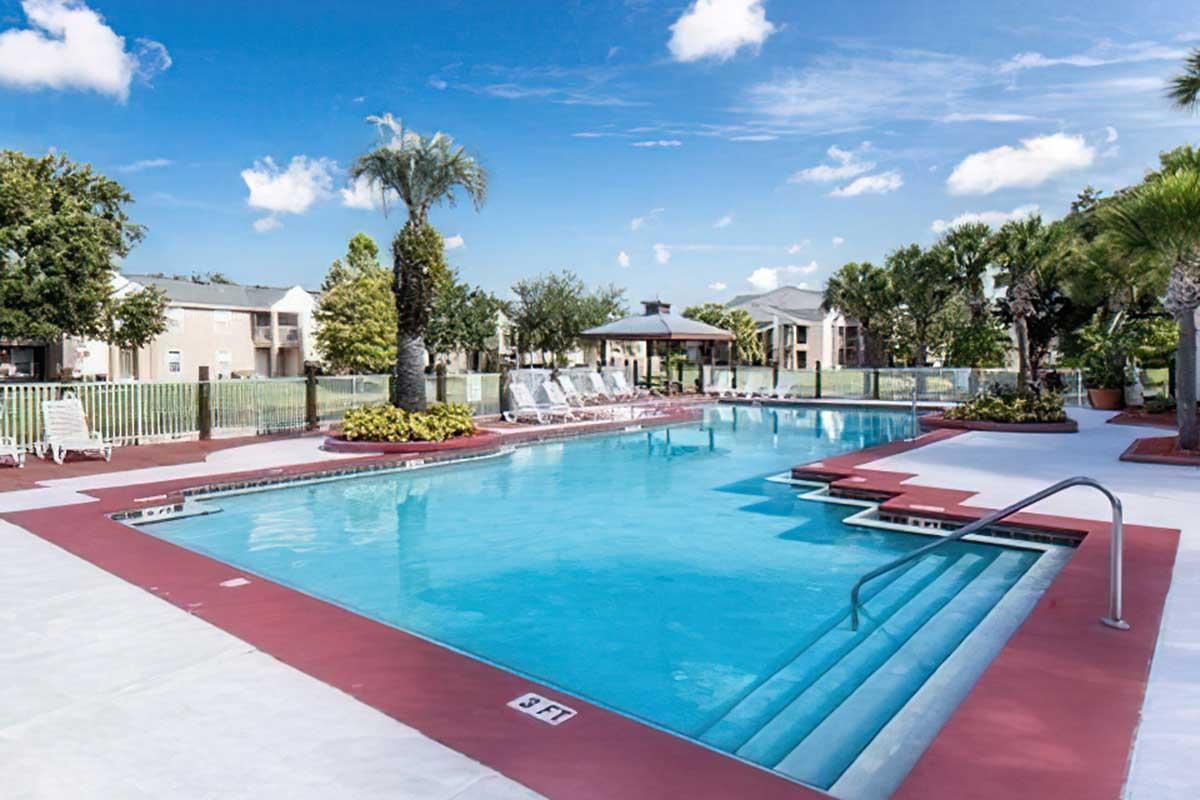 A clear blue swimming pool surrounded by a red deck, lounge chairs, and palm trees.