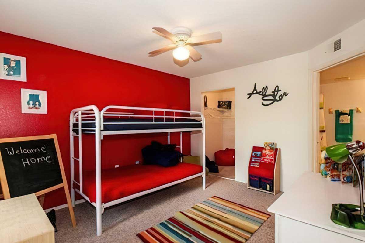 A cozy bedroom featuring a red accent wall, a white metal bunk bed with red bedding, a chalkboard welcoming guests, a colorful striped rug, and a small closet area.