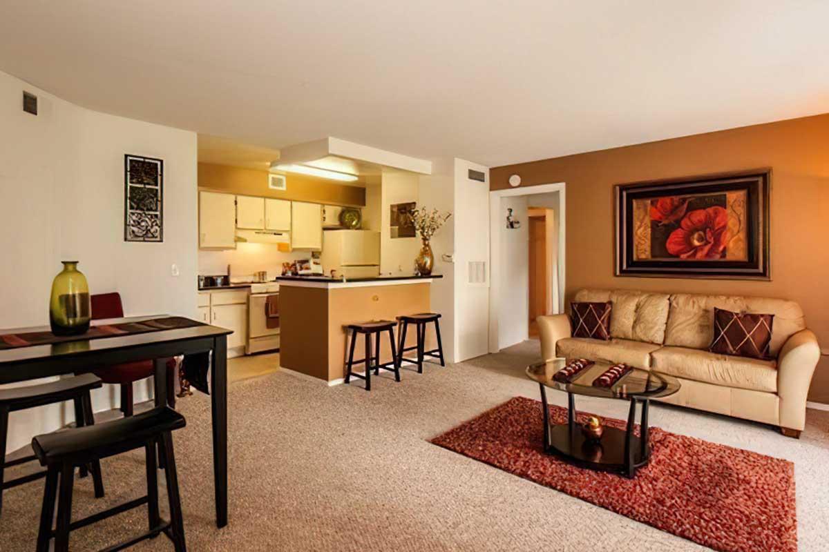 A cozy living room featuring a beige couch with red and brown accent pillows, a glass coffee table on a red rug, and a small dining area with a black table and stools.