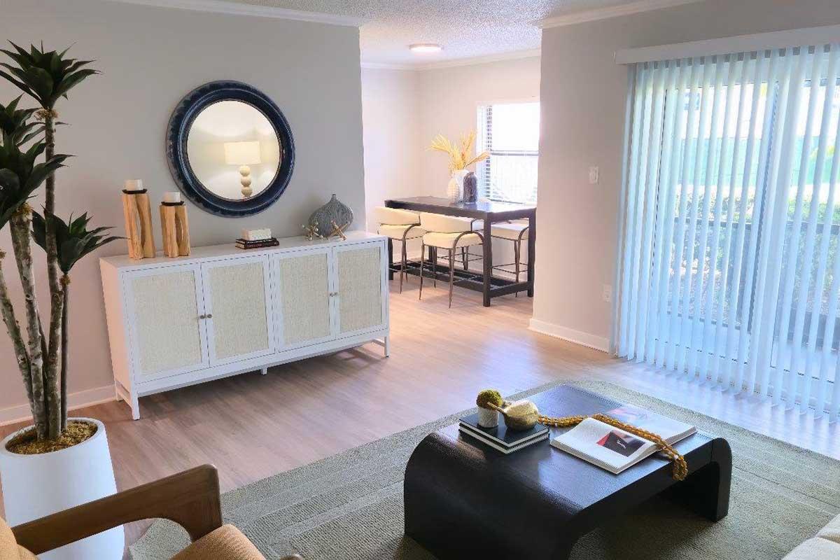 A modern living room featuring a neutral color palette with a wooden chair, a decorative plant, a white sideboard, and a round mirror. In the background, there's a dining area with bar stools and a window with vertical blinds letting in natural light.