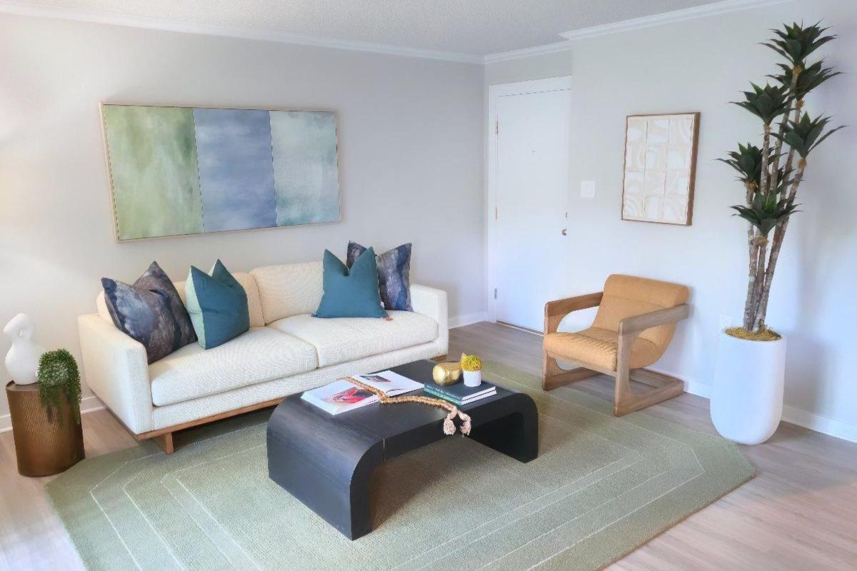 A modern living room featuring a light-colored sofa with teal cushions, a sculptural coffee table, and a decorative rug. A potted plant and a small cactus add greenery, while artwork on the walls provides a touch of color. Natural light fills the space, creating a welcoming and contemporary atmosphere.