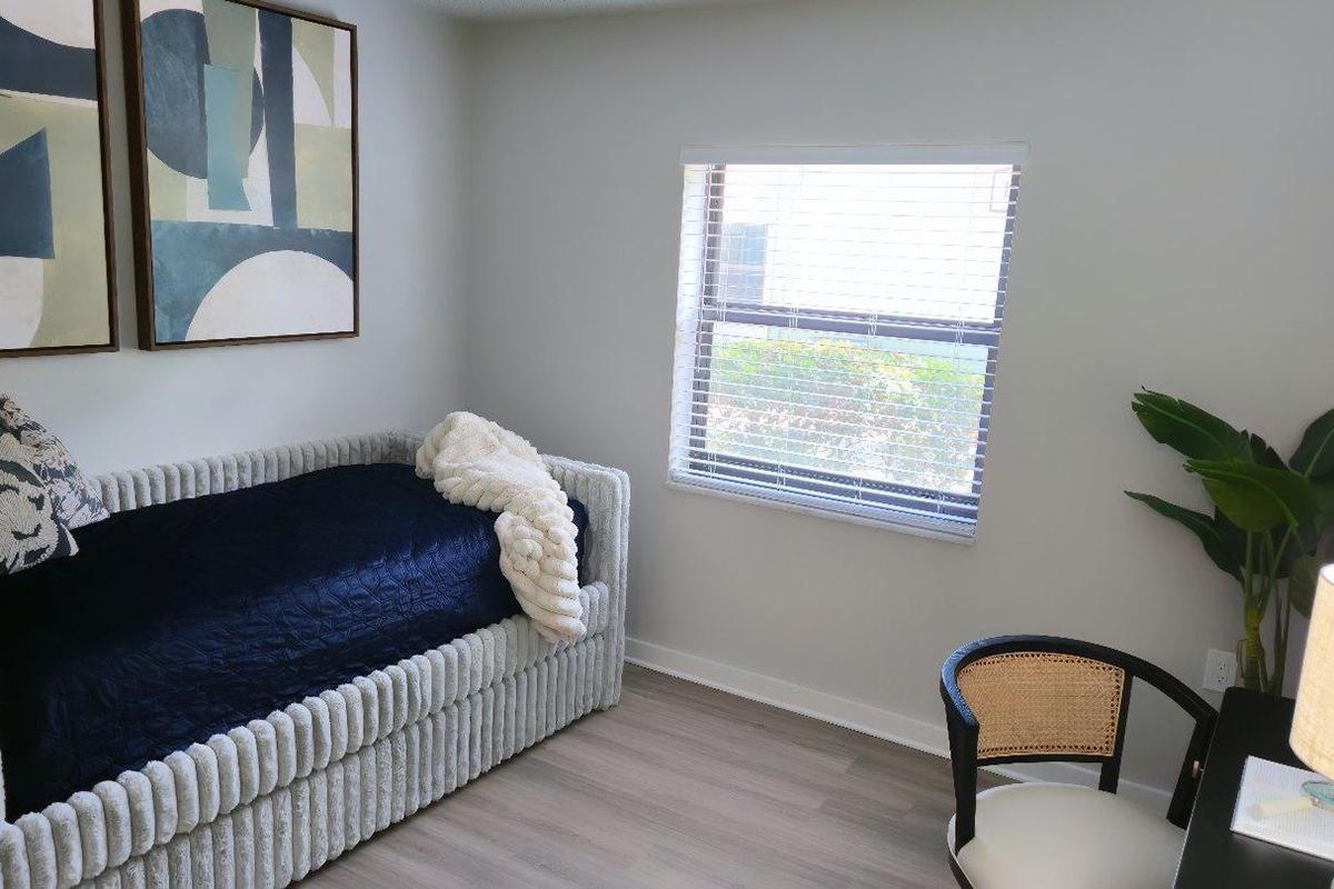 A cozy interior space featuring a textured, light-colored sofa adorned with a dark throw blanket and decorative pillows. A window with blinds provides natural light, revealing greenery outside. A black and natural woven chair sits beside a small table, adding to the inviting atmosphere of the room.