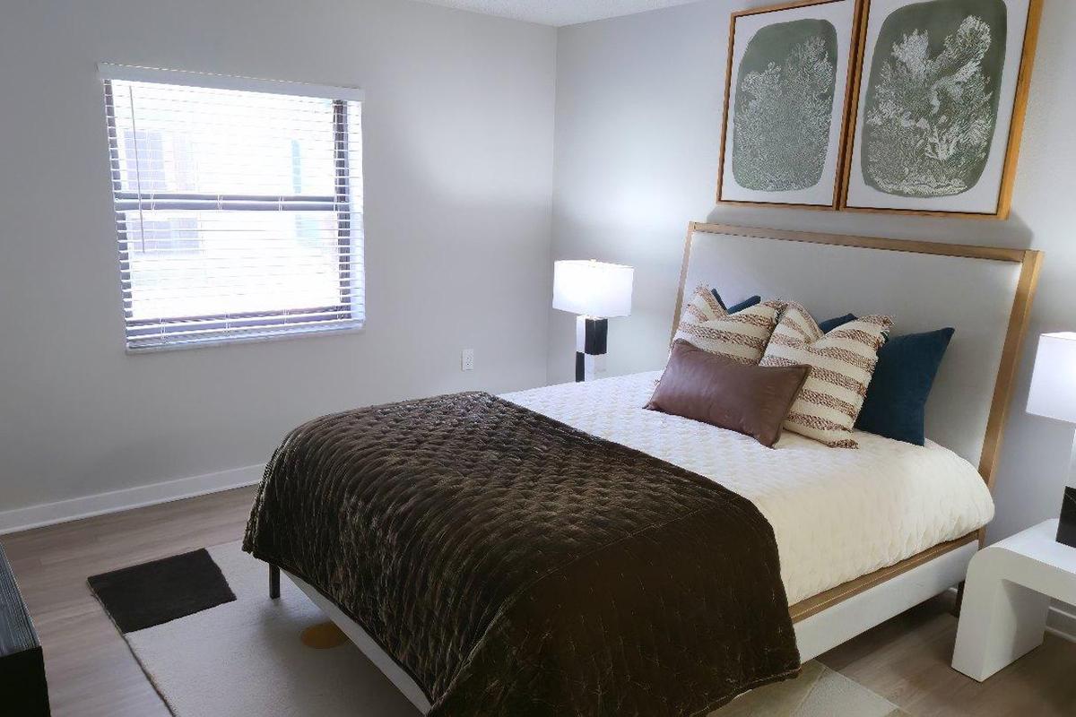A modern bedroom featuring a queen-sized bed with a dark brown throw blanket and decorative pillows. Two bedside lamps are on either side, and the walls are adorned with two framed botanical prints. A window with blinds allows natural light to enter, illuminating the cozy space with light-colored walls and a minimalist aesthetic.
