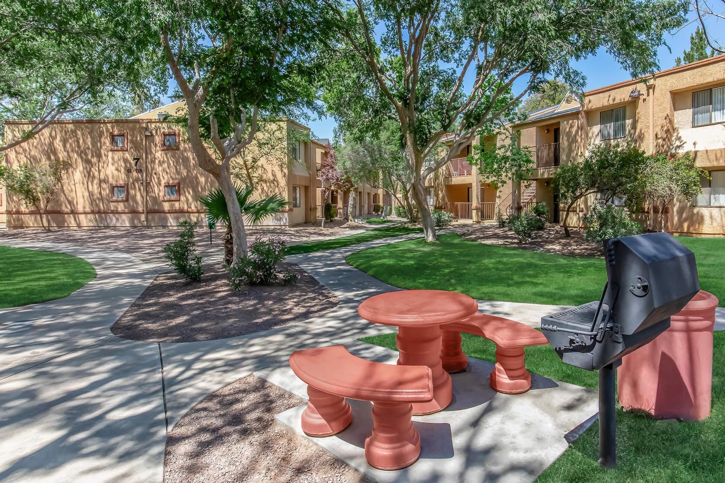 A landscaped courtyard featuring a red picnic table and grill surrounded by grass and trees. Two buildings with balconies are visible in the background, creating a welcoming outdoor space for gatherings and relaxation. Pathways meander through the area, enhancing the scenic setting.