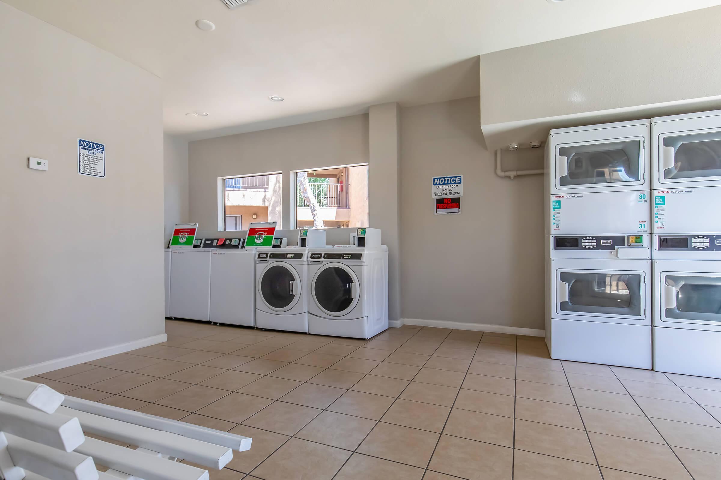 A clean and modern laundry room featuring several washing machines and dryers against a light-colored wall. There are windows letting in natural light and a bench for seating. The tiled floor and minimal decorations create a functional and inviting space for doing laundry.