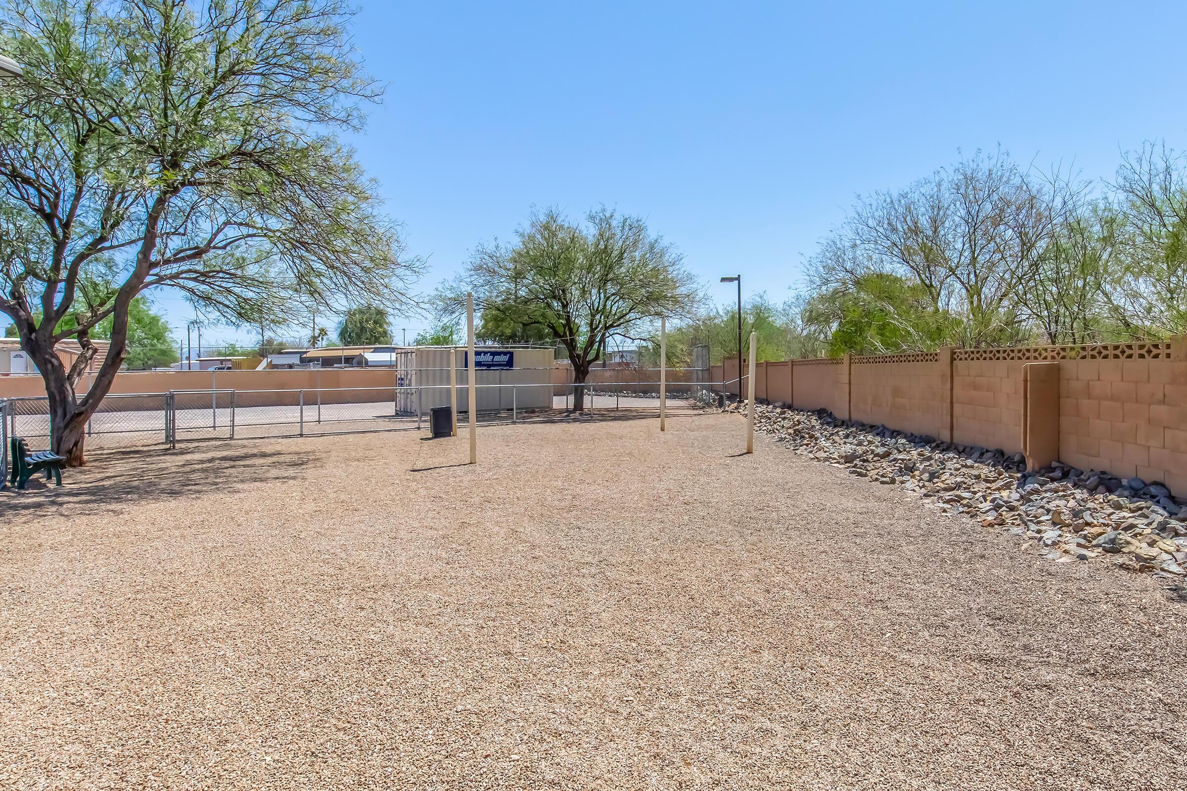 A spacious outdoor area featuring a gravel surface, surrounded by low walls and desert vegetation. There are a couple of trees, a play structure in the distance, and a few light poles. The sky is clear and blue, indicating a sunny day.