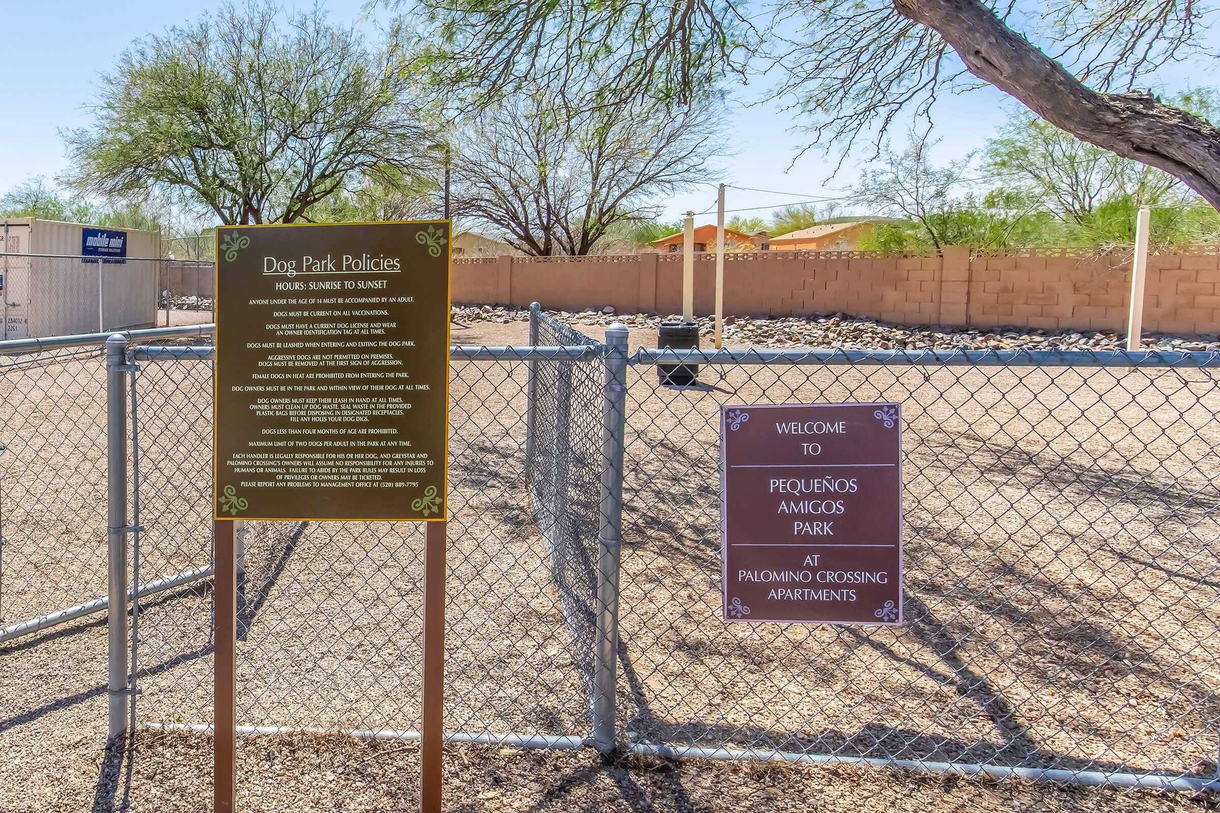 Signage at the entrance of a dog park, detailing park policies on one side and a welcome message to "Pequeños Amigos Park at Palomino Crossing Apartments" on the other, surrounded by a chain-link fence and sparse landscaping.