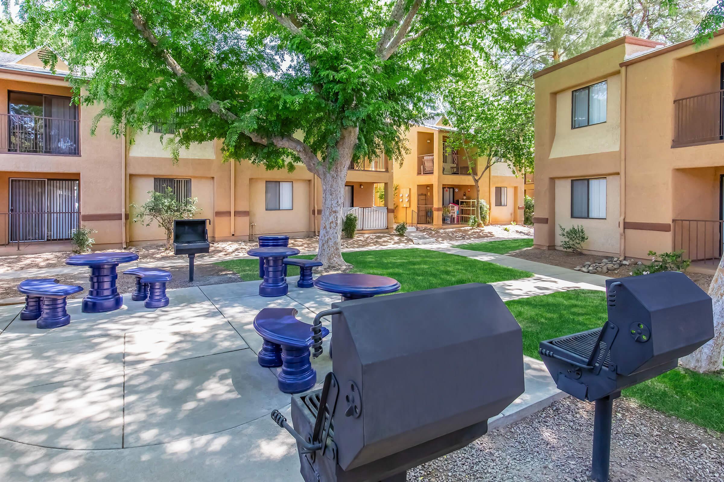 Outdoor courtyard area featuring two charcoal grills, picnic tables with vibrant blue seats, and well-maintained green grass. Surrounding the space are tan-colored apartment buildings with balconies. The setting is shaded by large trees, creating a welcoming environment for gatherings.