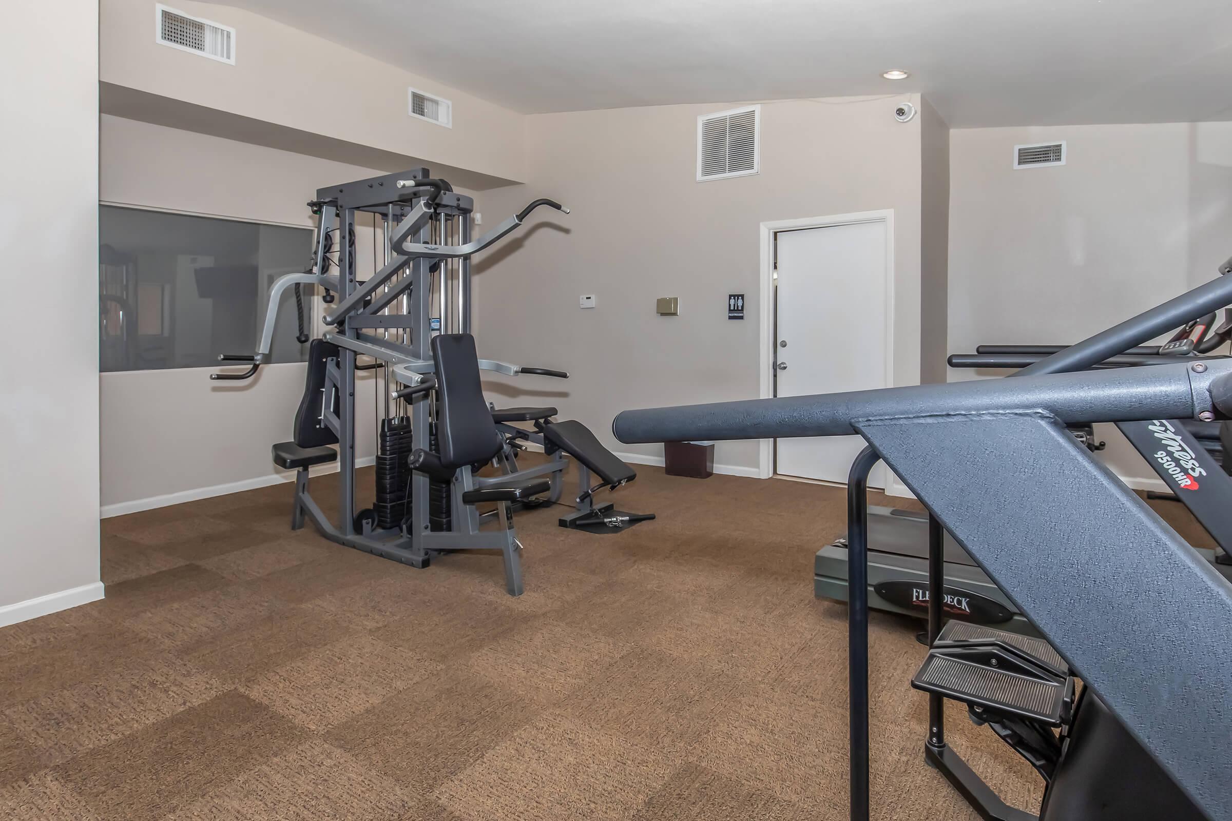 A spacious fitness room featuring various exercise equipment, including a multi-gym setup, weight bench, and treadmills. The walls are a neutral color, with carpeting on the floor. Large windows provide natural light, and there is a door leading outside.