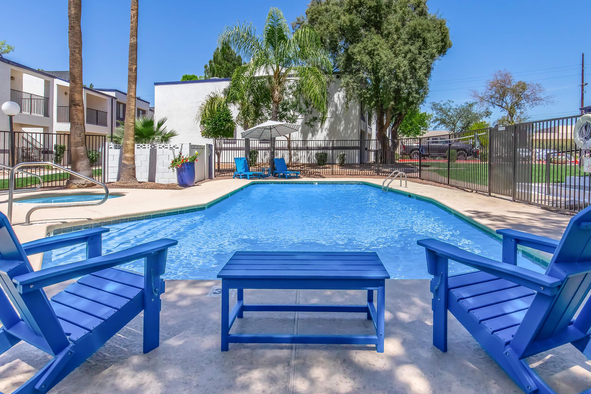 A clear blue swimming pool surrounded by palm trees and chairs. Two blue lounge chairs face the pool, with a small table between them. In the background, there are trees, a fence, and several umbrellas providing shade. Bright blue sky complements the sunny atmosphere.